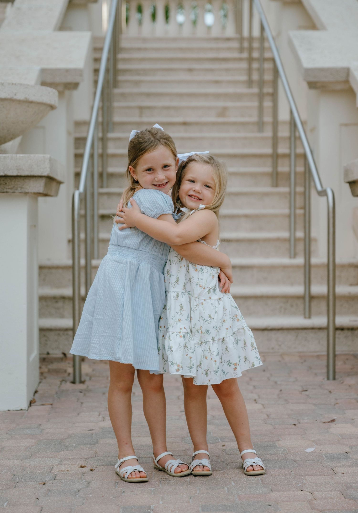 Two young sisters in summer dresses share a warm hug in front of an outdoor staircase.