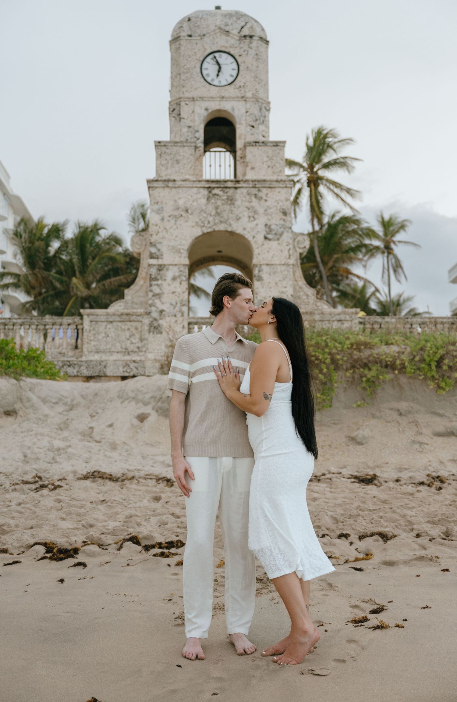 Couple sharing a kiss on the beach in front of a historic clock tower with palm trees in Palm Beach.