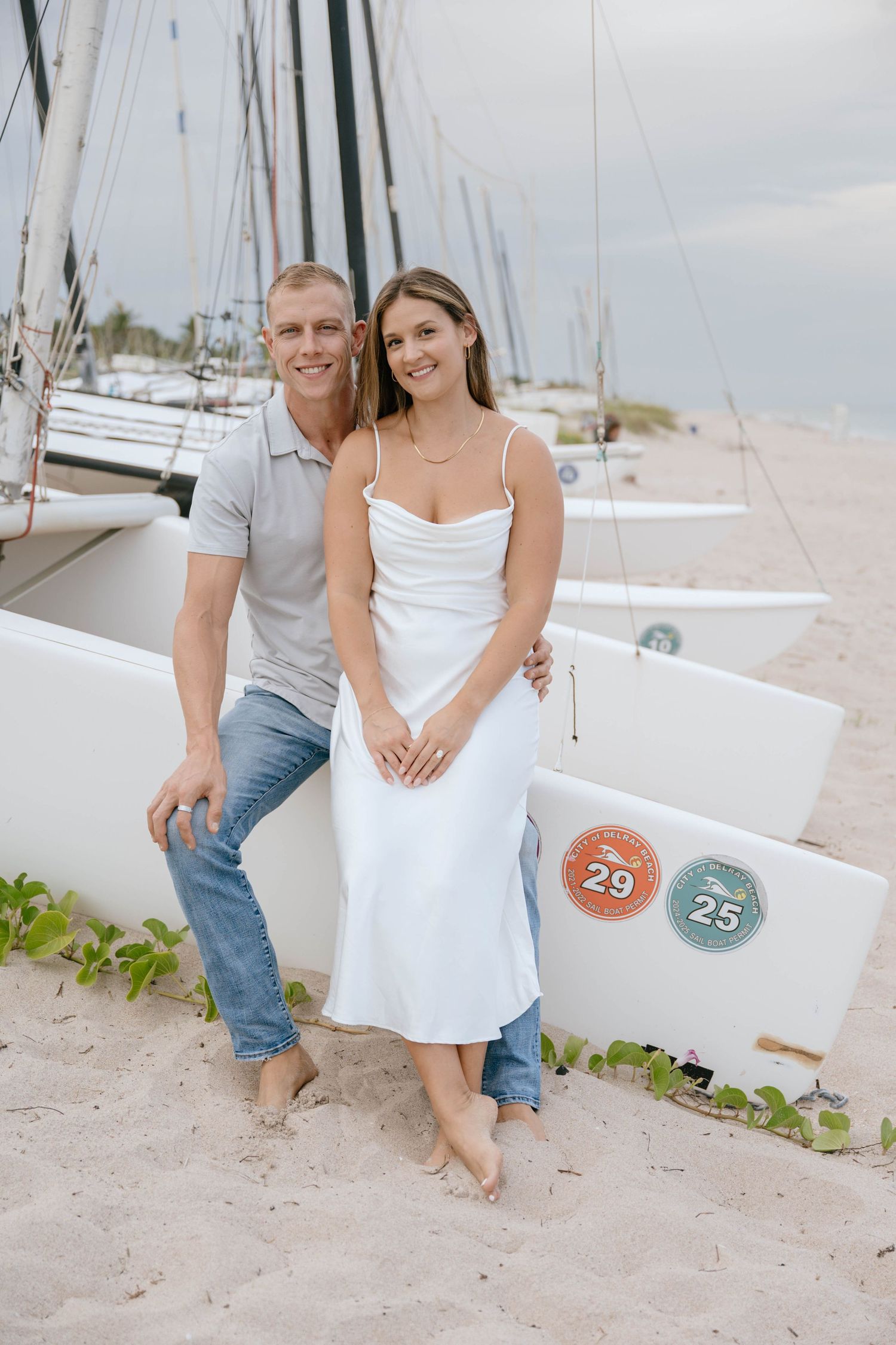 Smiling engaged couple in casual attire leaning against sailboats numbered 25 and 29 on sandy beach.