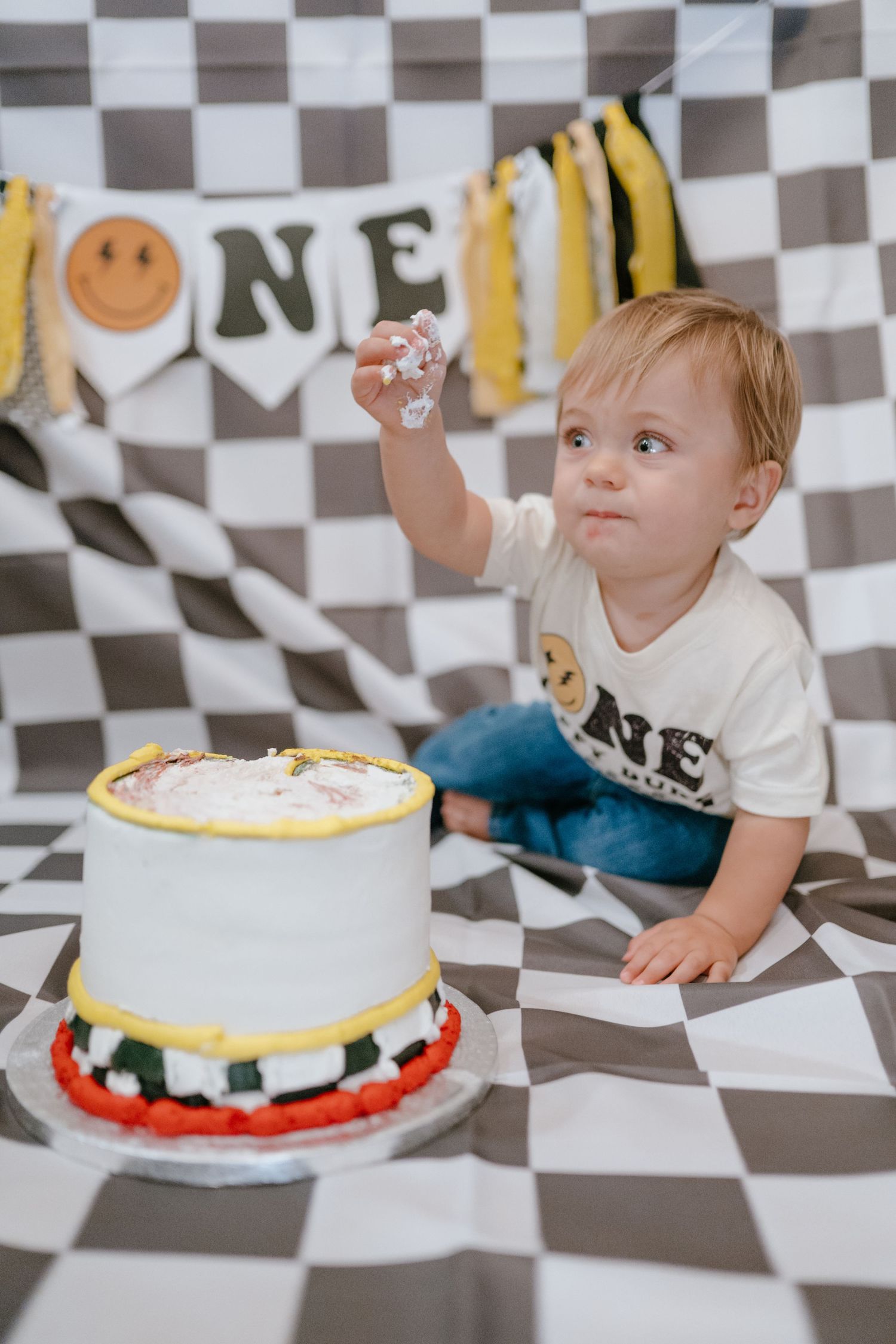 Toddler in ONE shirt reaches toward his decorated first birthday smash cake on checkered backdrop.