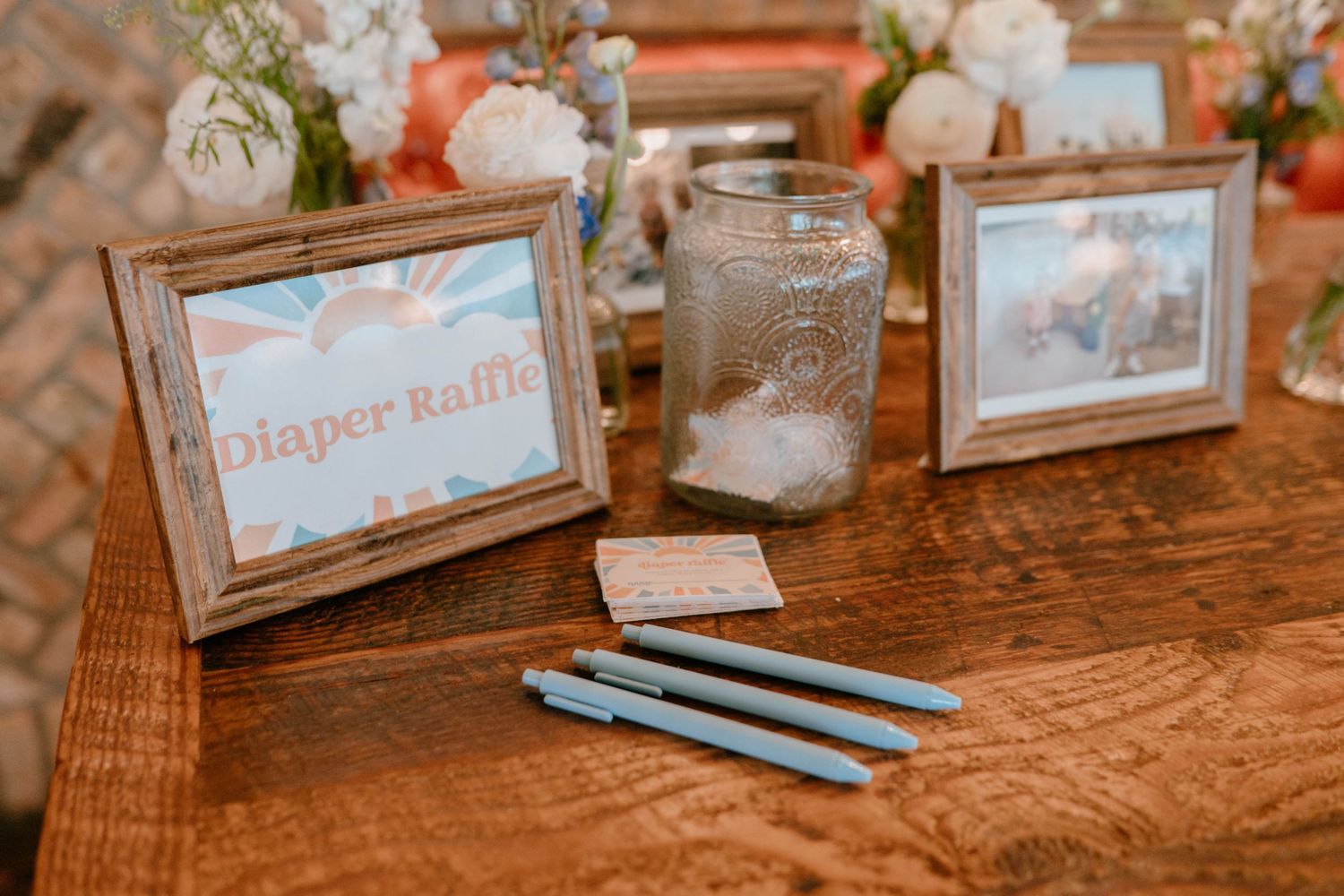Baby shower diaper raffle station with framed sign, blue pens, and mason jar centerpiece on wooden table.