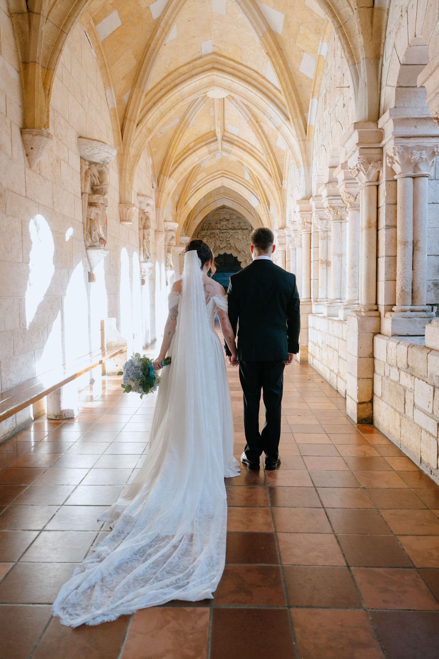 Bride and groom walking away hand in hand through sunlit Gothic stone cloister with long veil trailing.