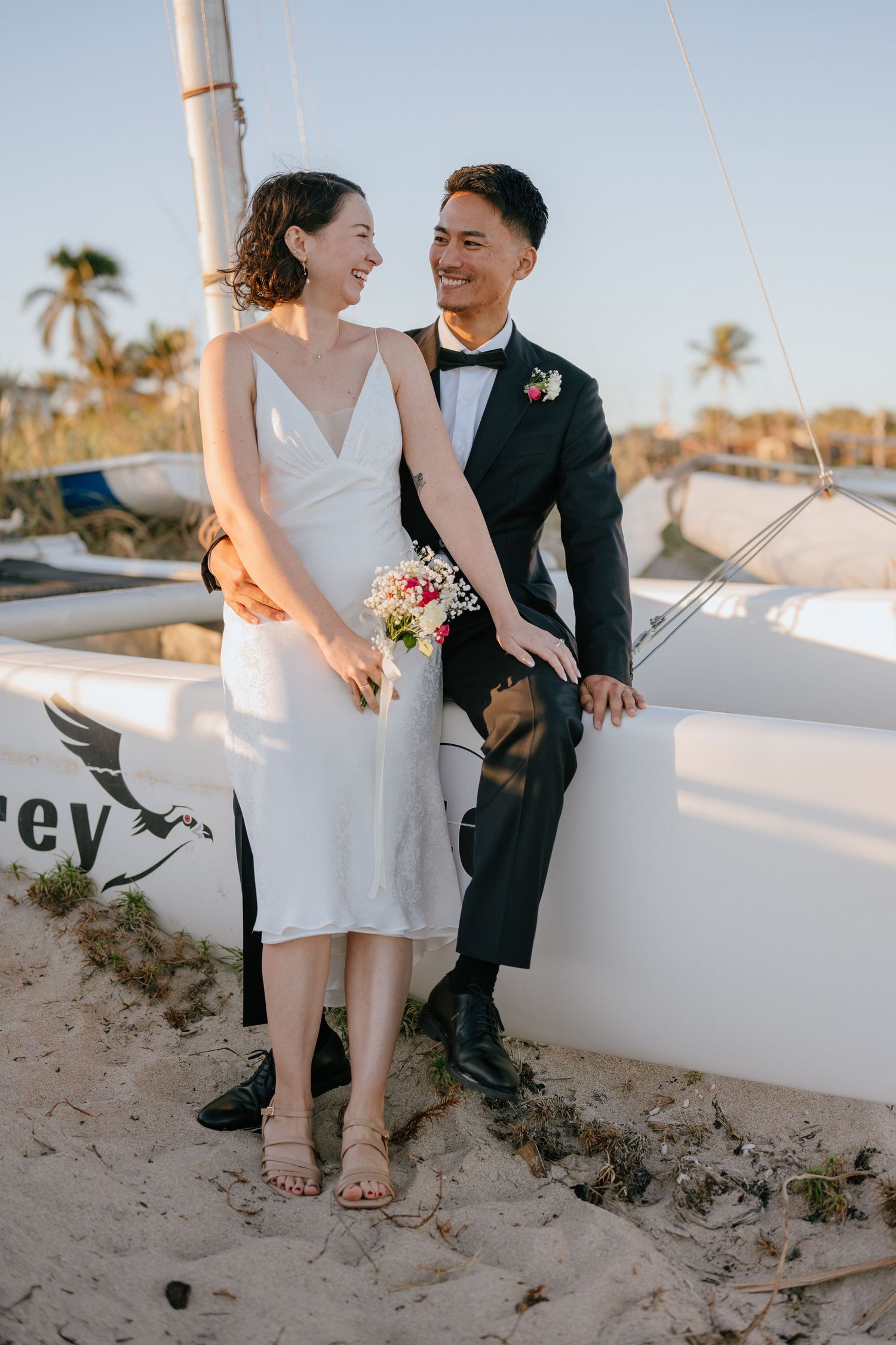 Smiling couple on a sandy beach near a sailboat, bride in white slip dress holding a bouquet.