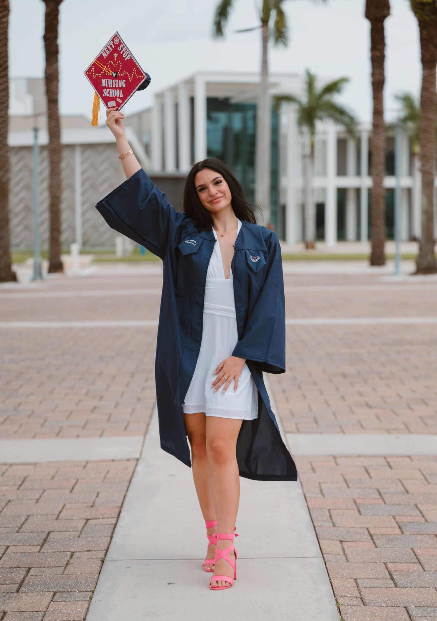 FAU graduate in navy gown raises decorated nursing school cap beside palm trees on campus.