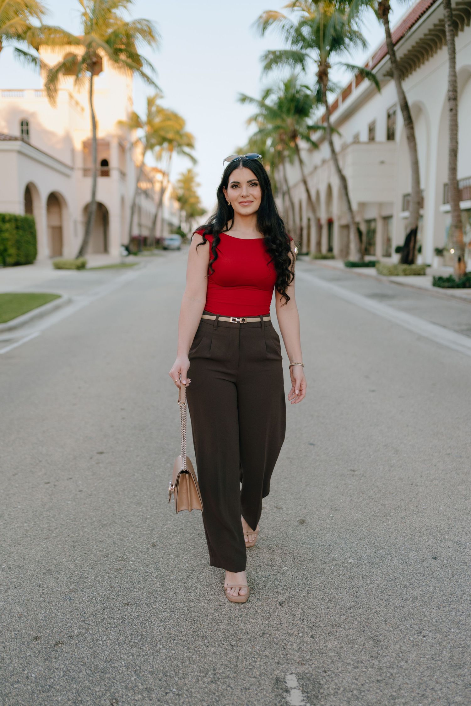 Woman in red top and brown trousers walking palm-lined street holding beige bag.