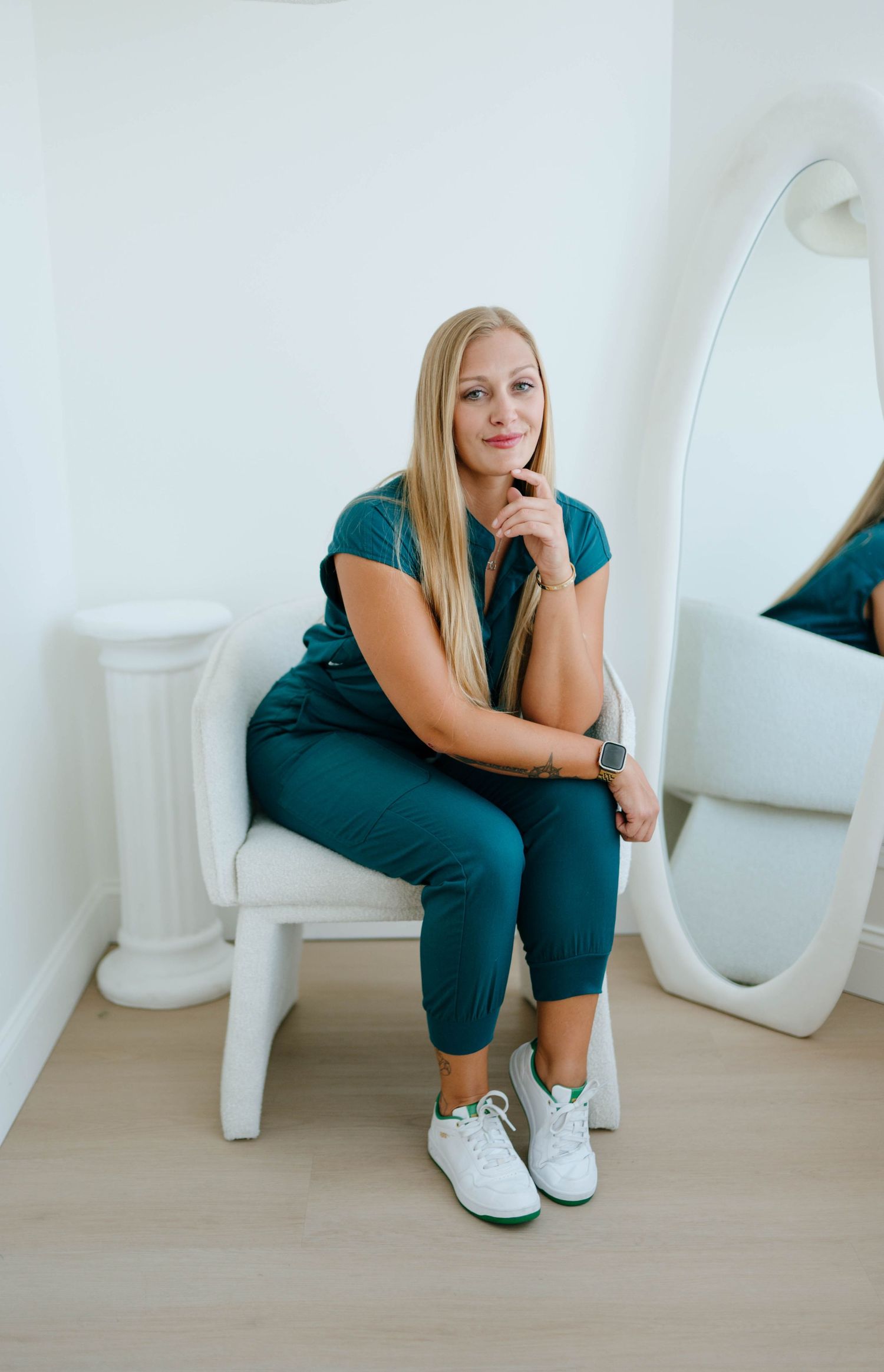 Blonde woman in teal medical scrubs sitting on a white chair beside a large oval mirror in a bright room.