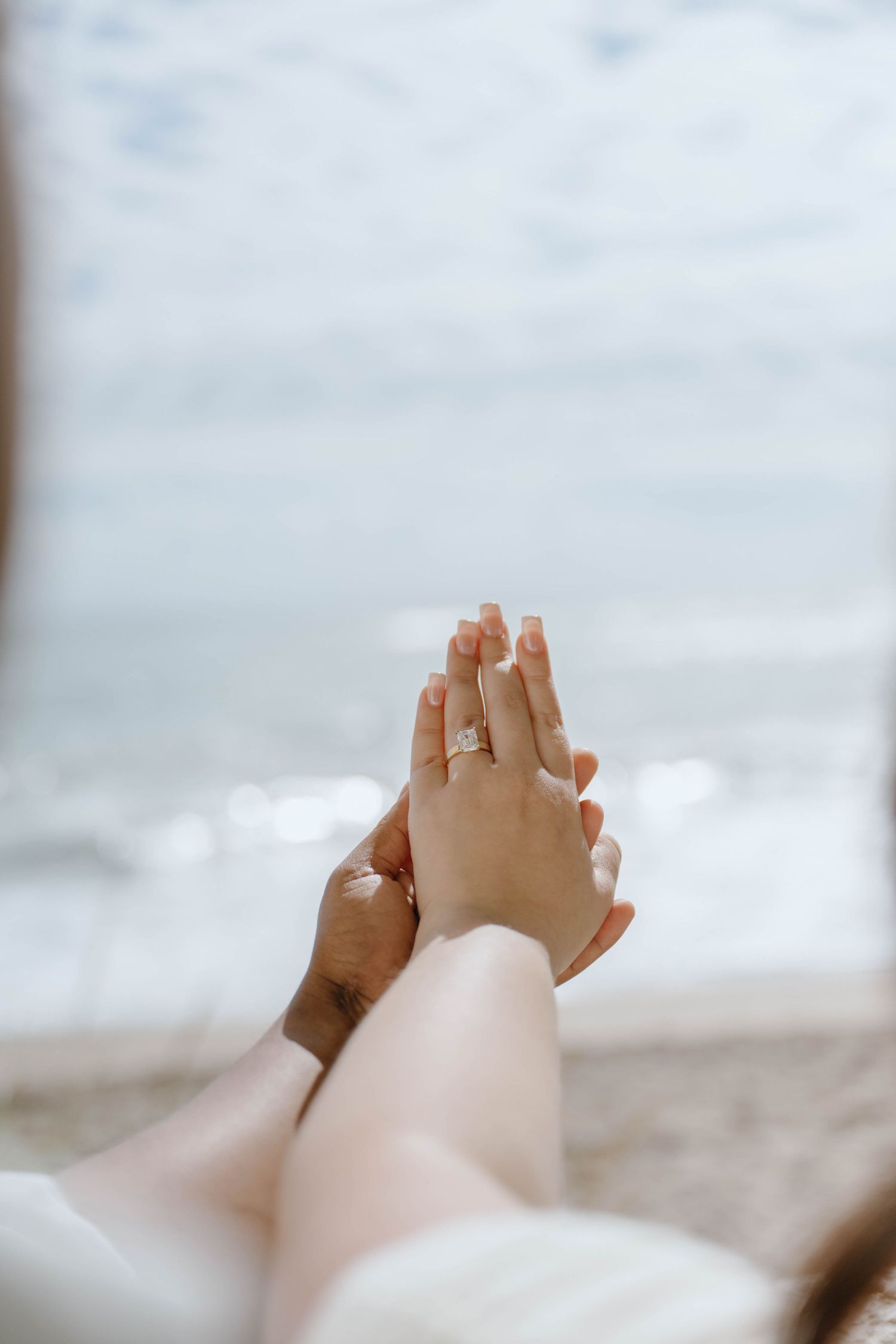 Close-up of engaged couple's hands pressed together showcasing a diamond engagement ring by the sea.