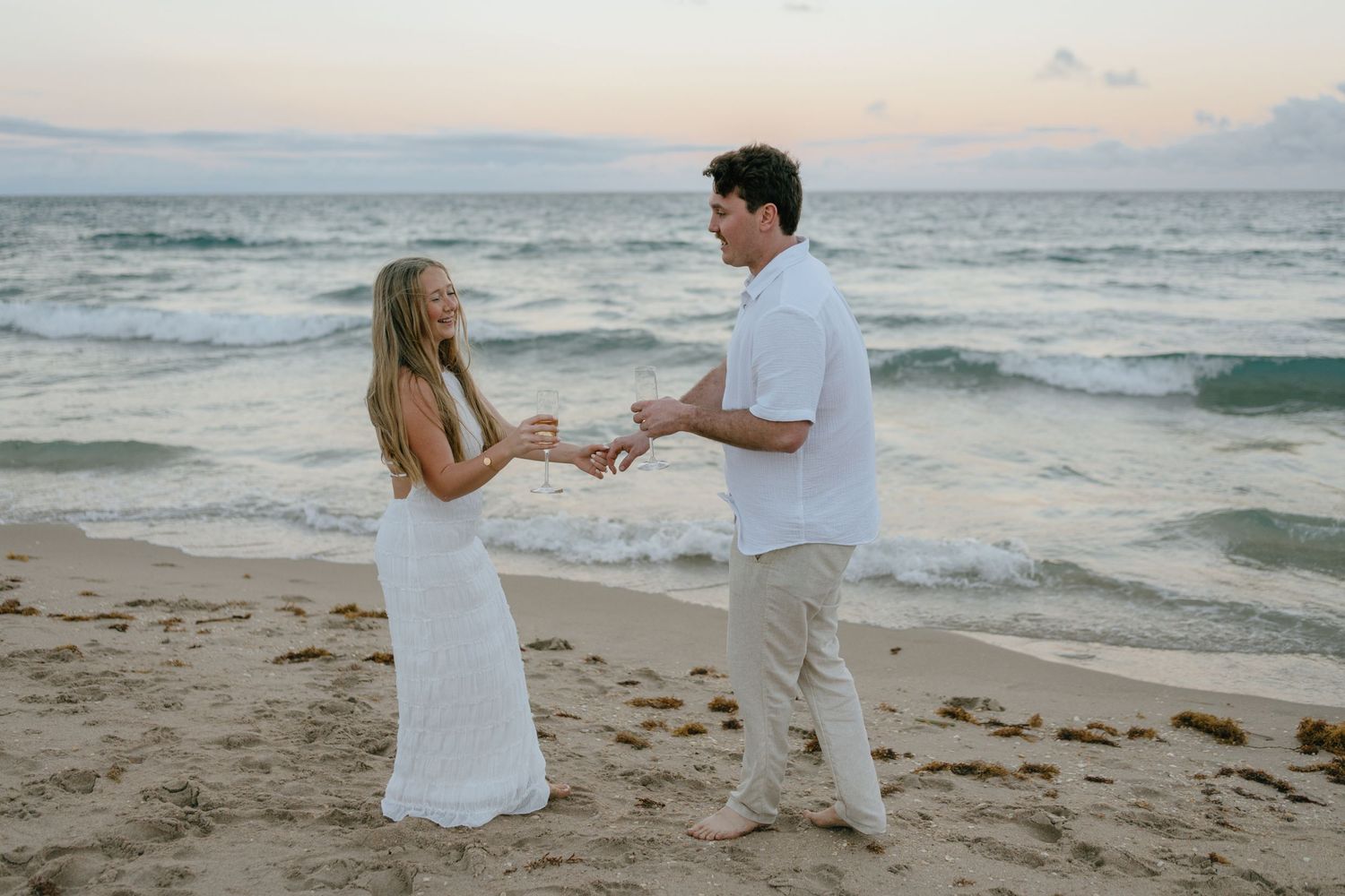 Engaged couple holding hands and smiling at each other on a sandy beach during a golden sunset session.