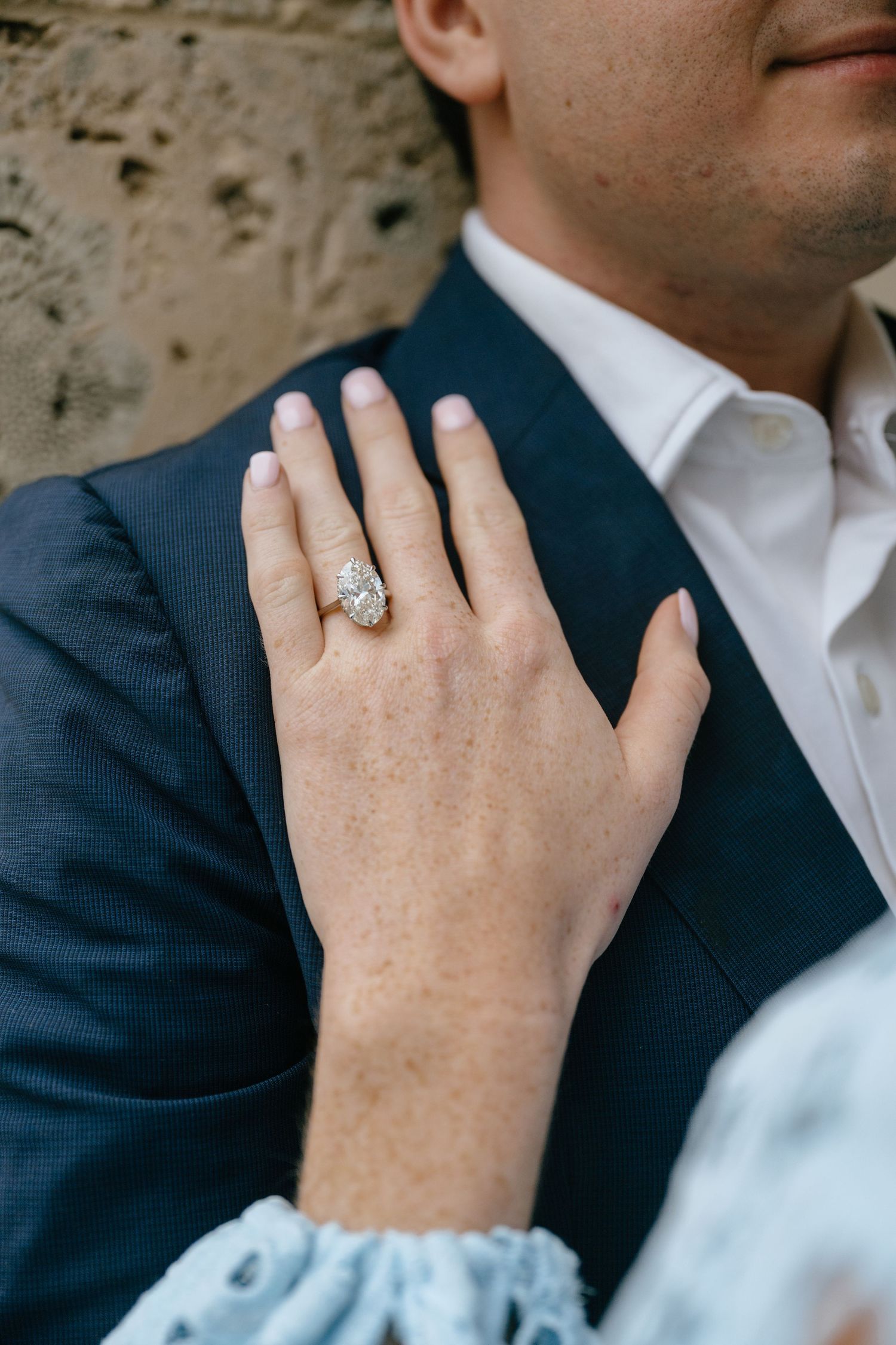 Close-up of a woman's hand displaying a stunning diamond engagement ring resting on a man's navy blue suit jacket.