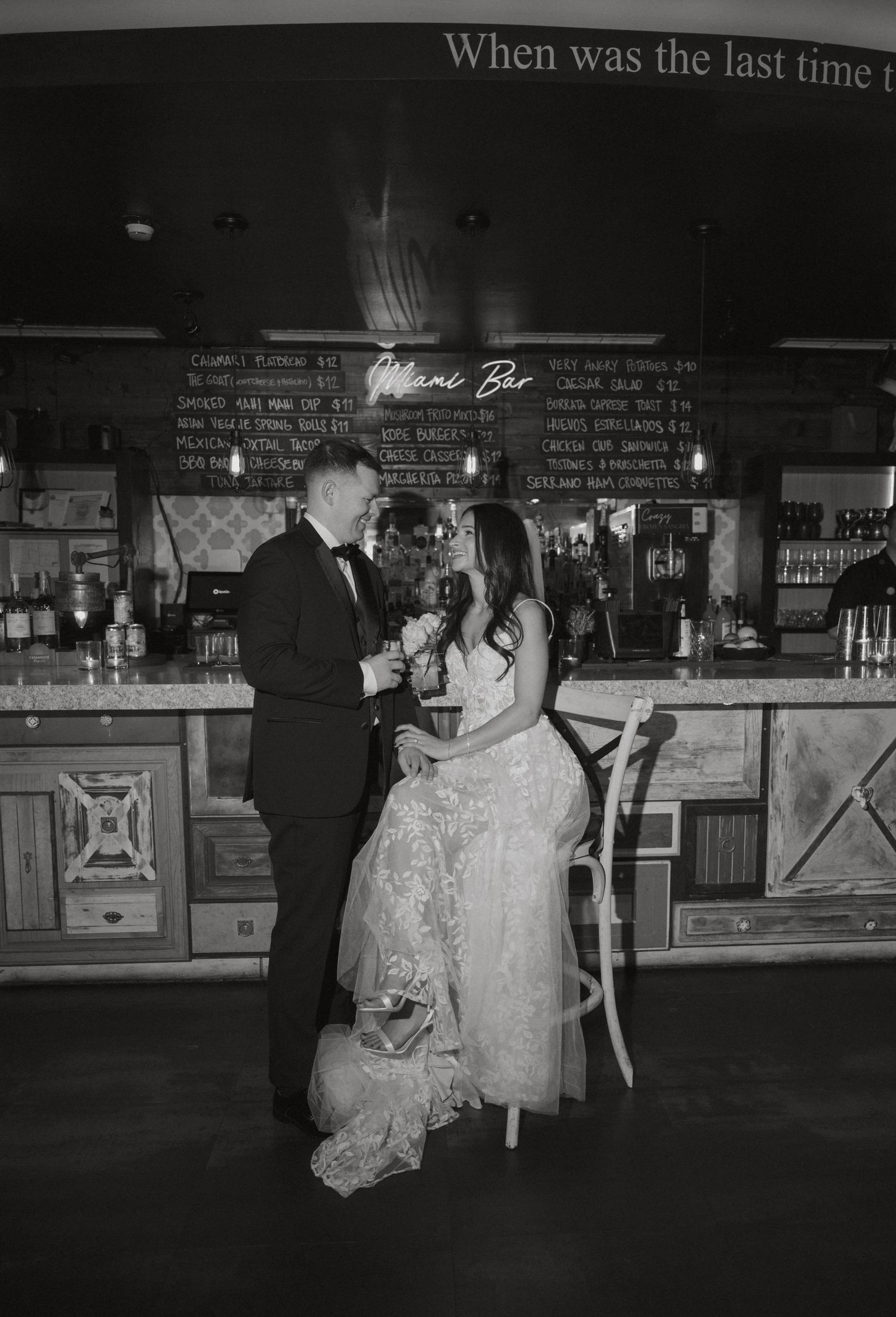 Black and white photo of bride and groom sharing a drink together at a rustic wedding bar.