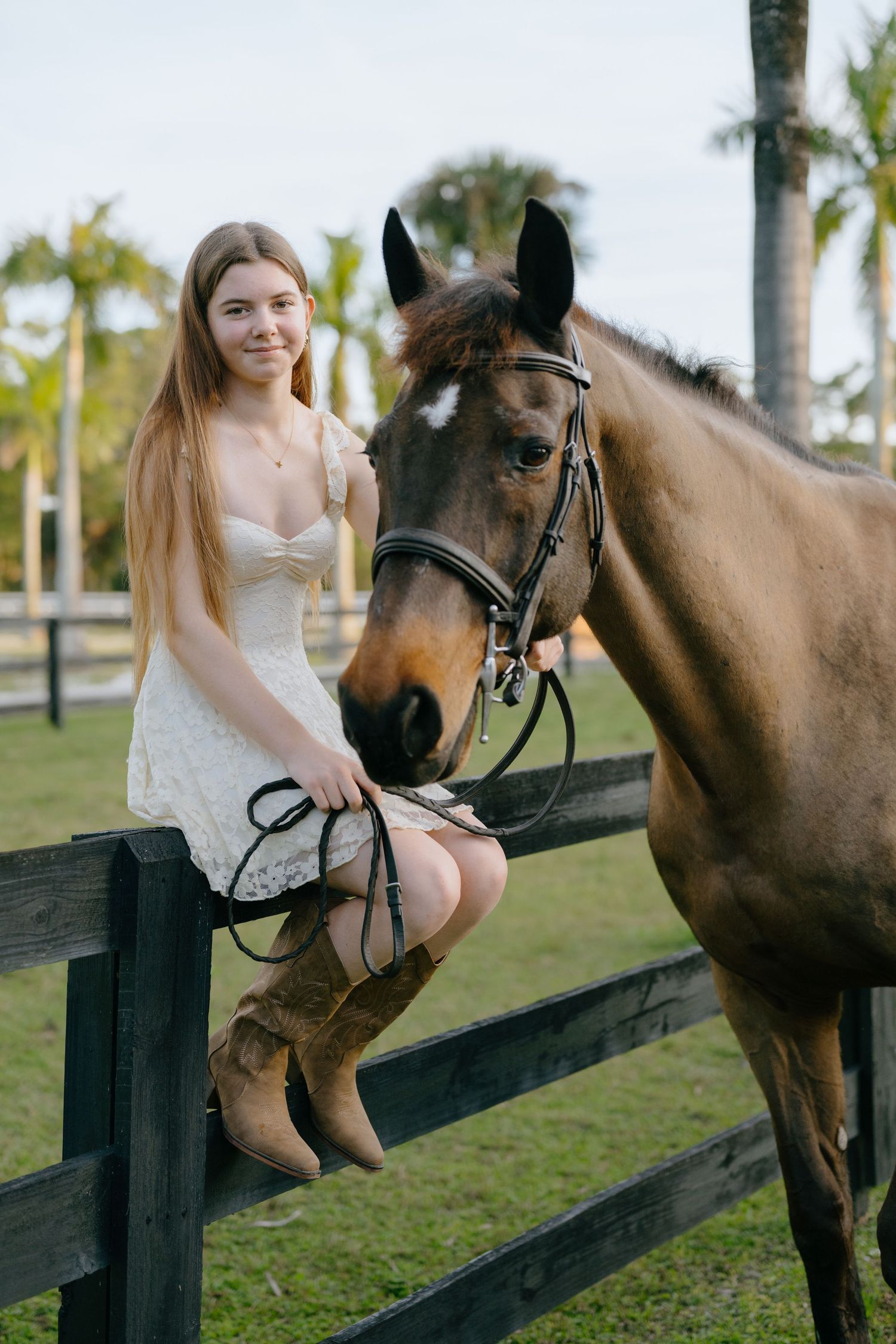 Young woman in white dress and cowboy boots sitting on fence beside brown horse with palm trees.