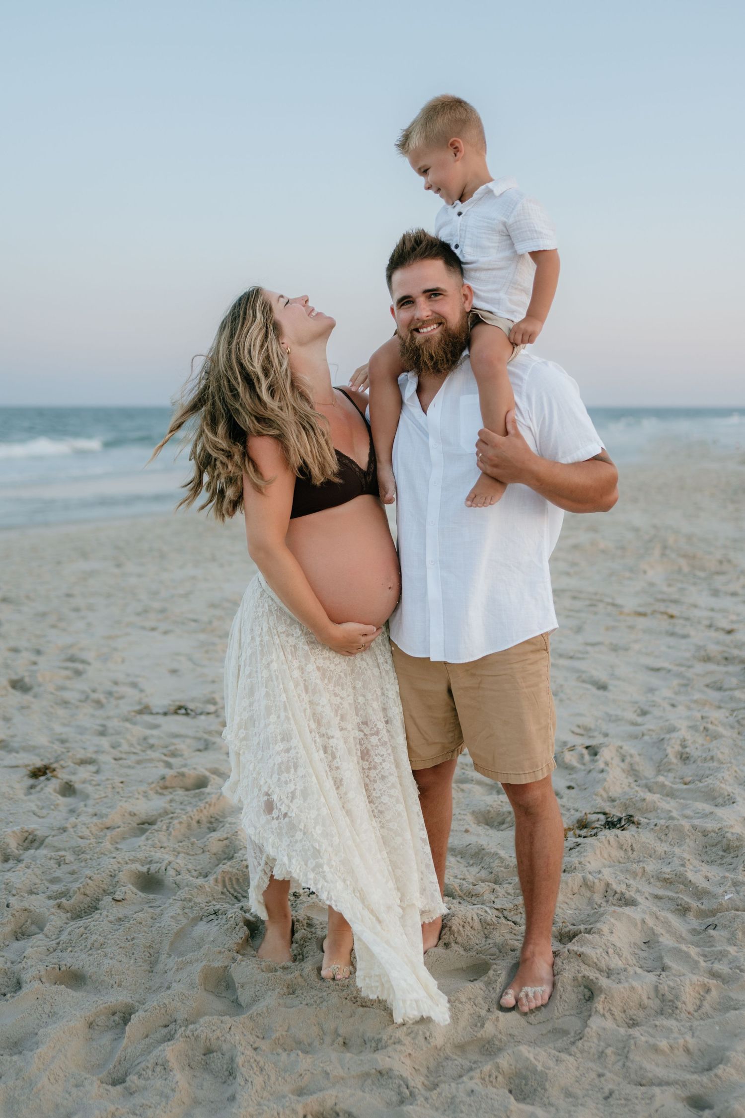 Joyful pregnant woman and bearded man with young boy on his shoulders at beach.