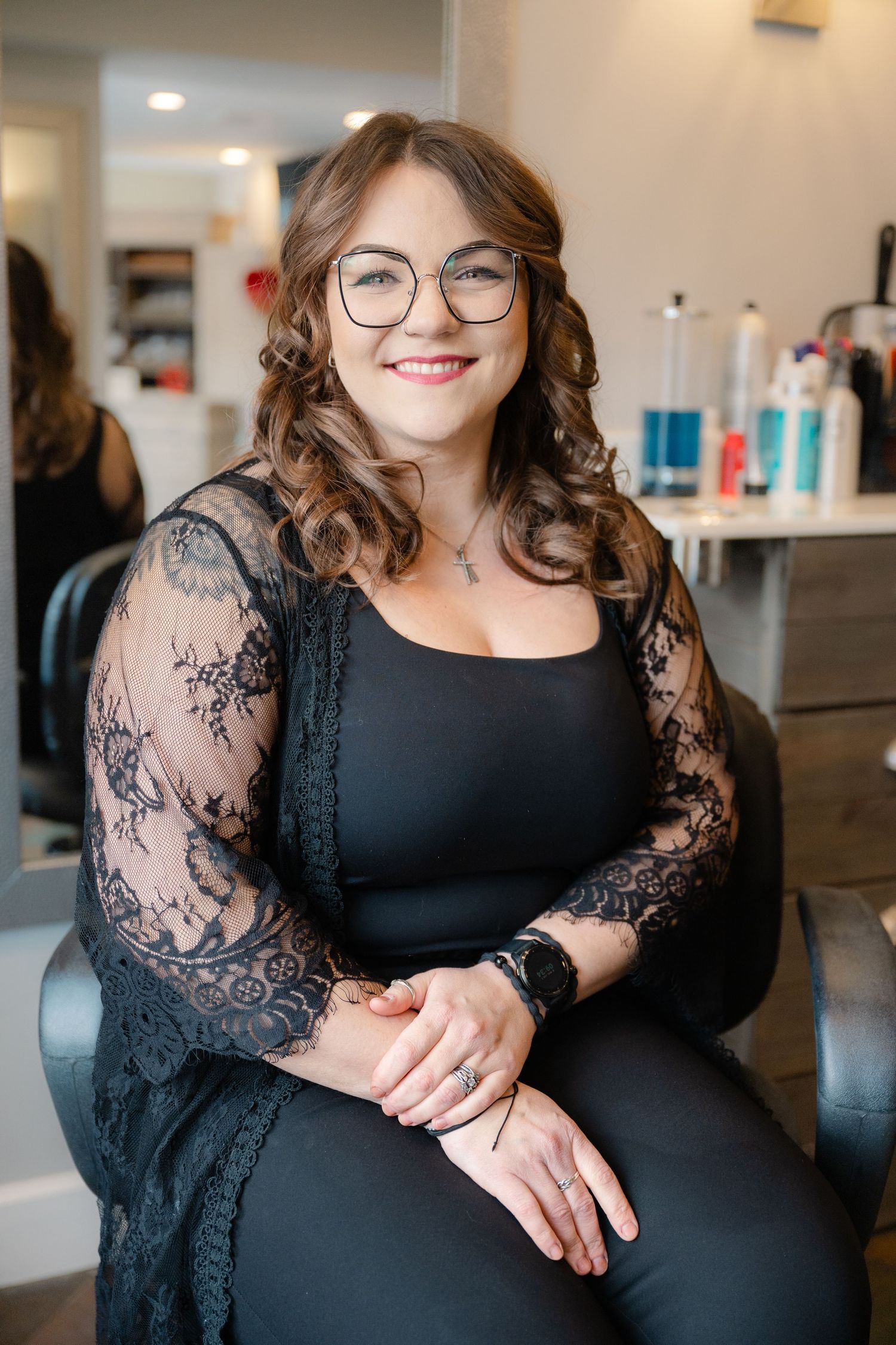 Smiling female hair stylist with glasses and curly brown hair sits in a salon chair wearing a black lace overlay top.