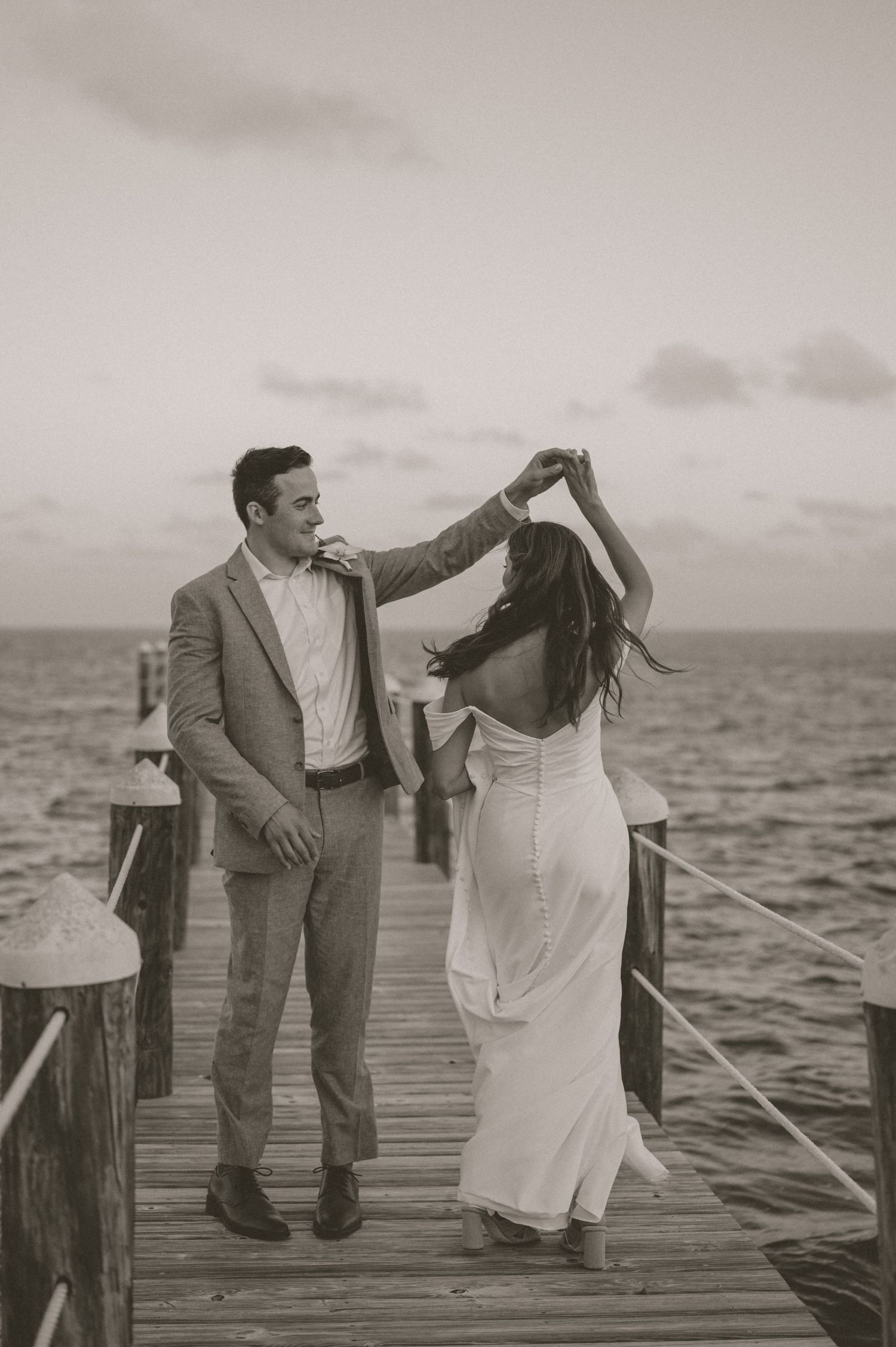 Black and white photo of groom spinning bride on a seaside dock at dusk, bride in a sleek backless gown.