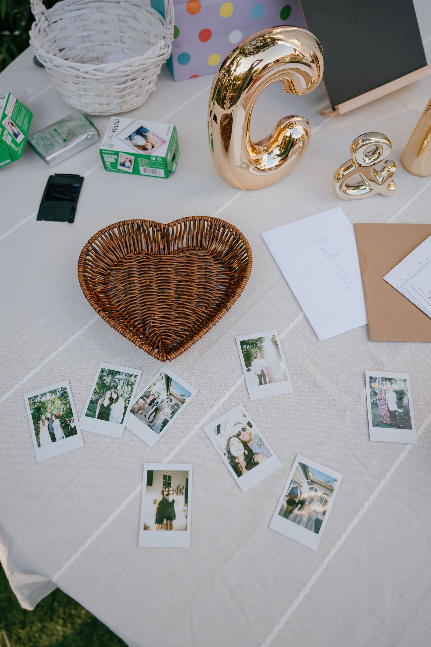 Polaroid photos scattered on a white tablecloth beside a heart-shaped wicker basket and gold letter decorations at a wedding reception.