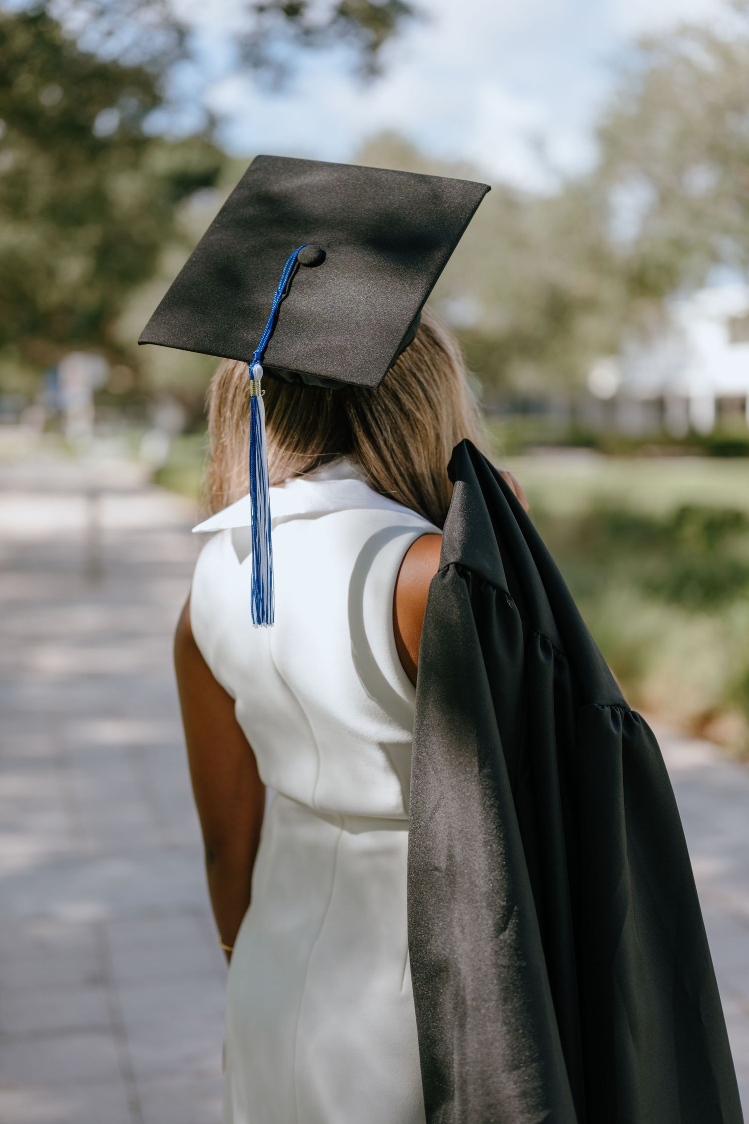 Back view of graduate in white dress wearing black mortarboard cap holding gown outdoors.