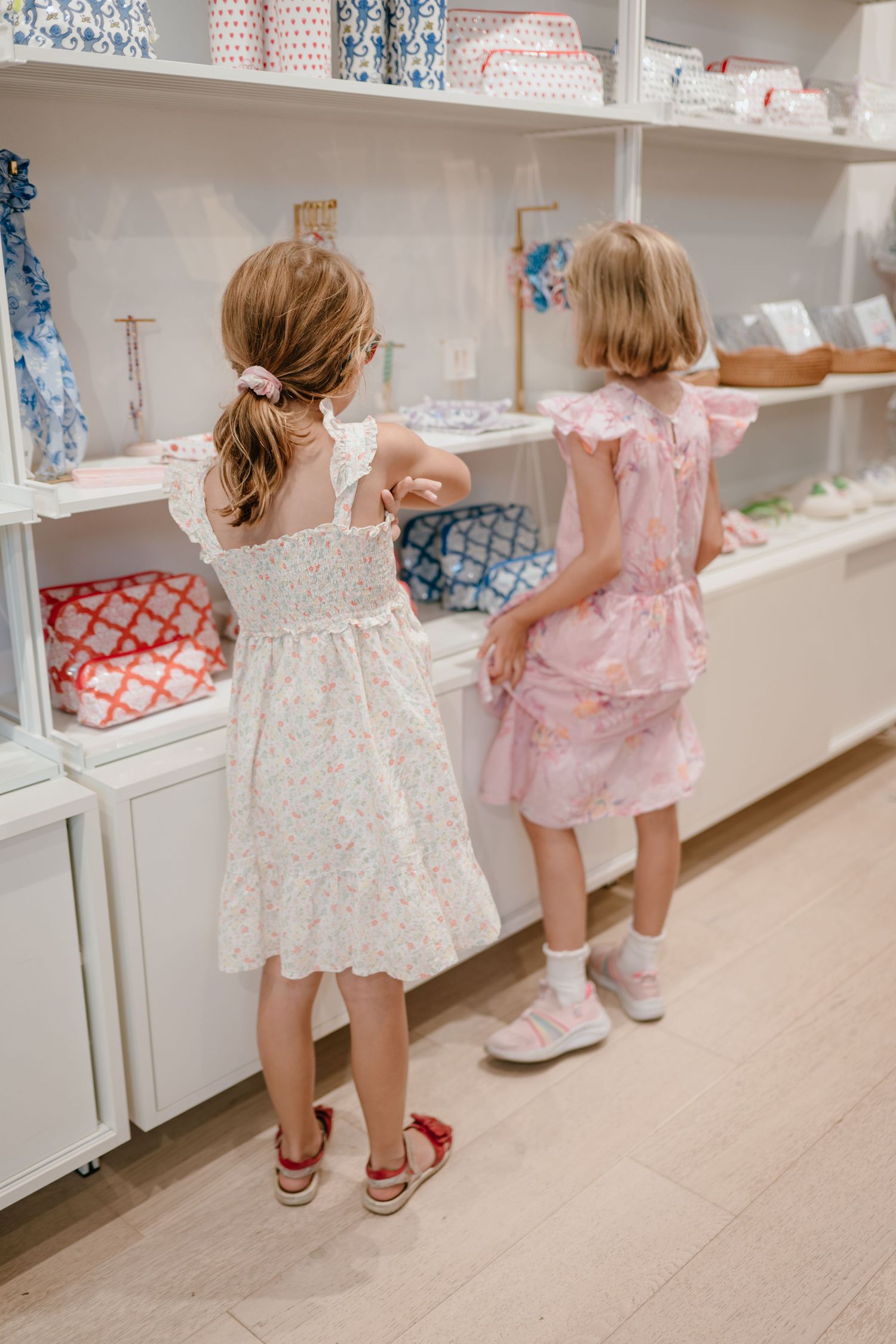 Two young girls in floral dresses browse colorful items on white retail shelves in a boutique store.