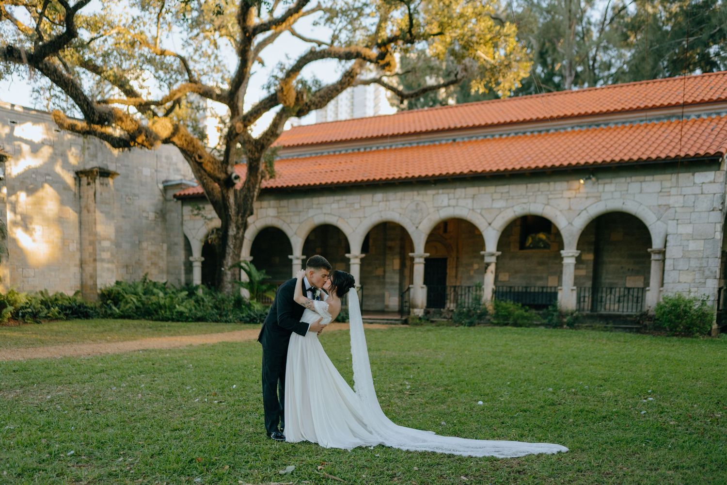 Bride and groom embracing on green lawn outside Spanish-style building with red tile roof at golden hour.