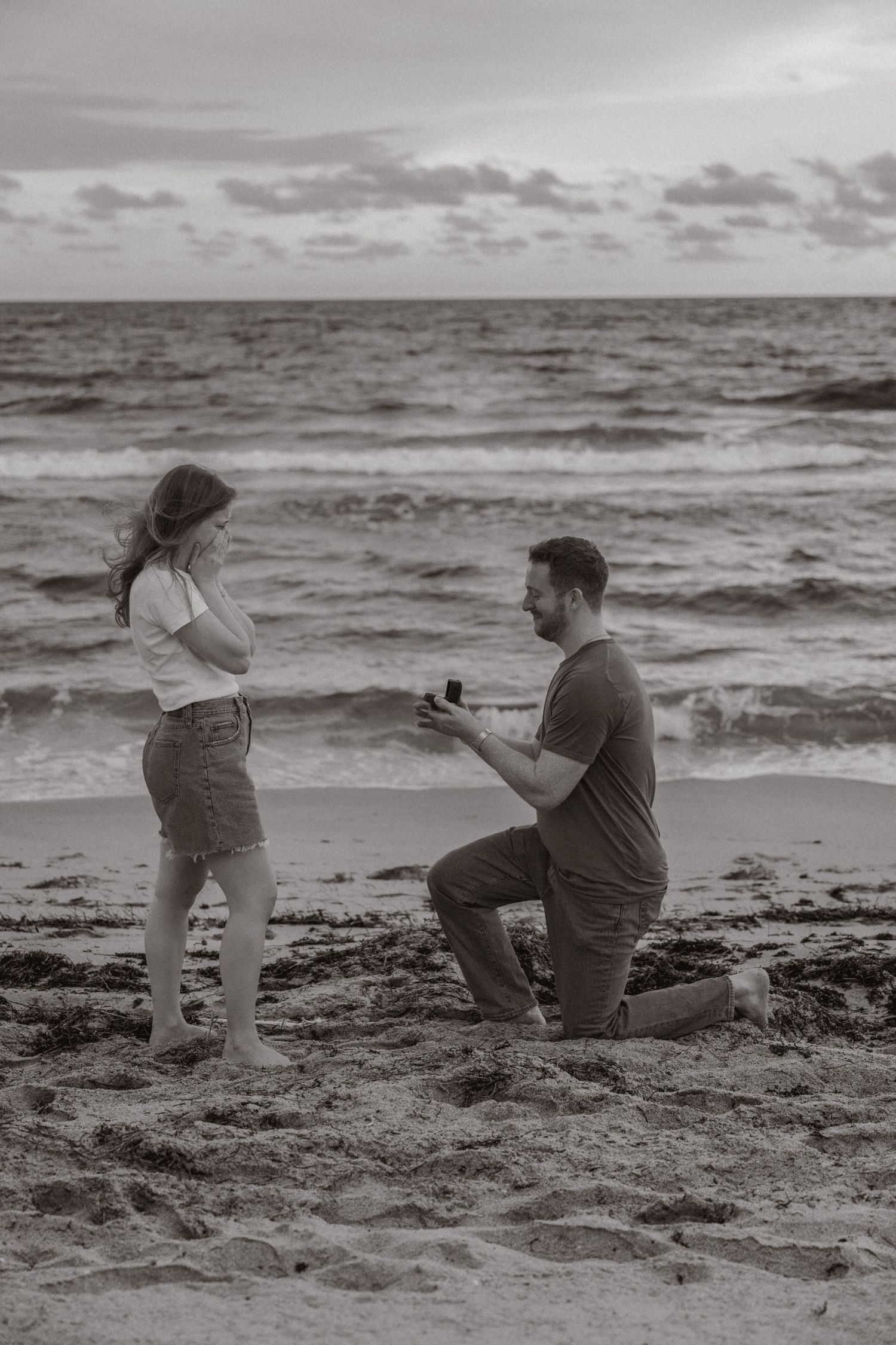 Man proposing on one knee to woman on beach at dusk, black and white photo.