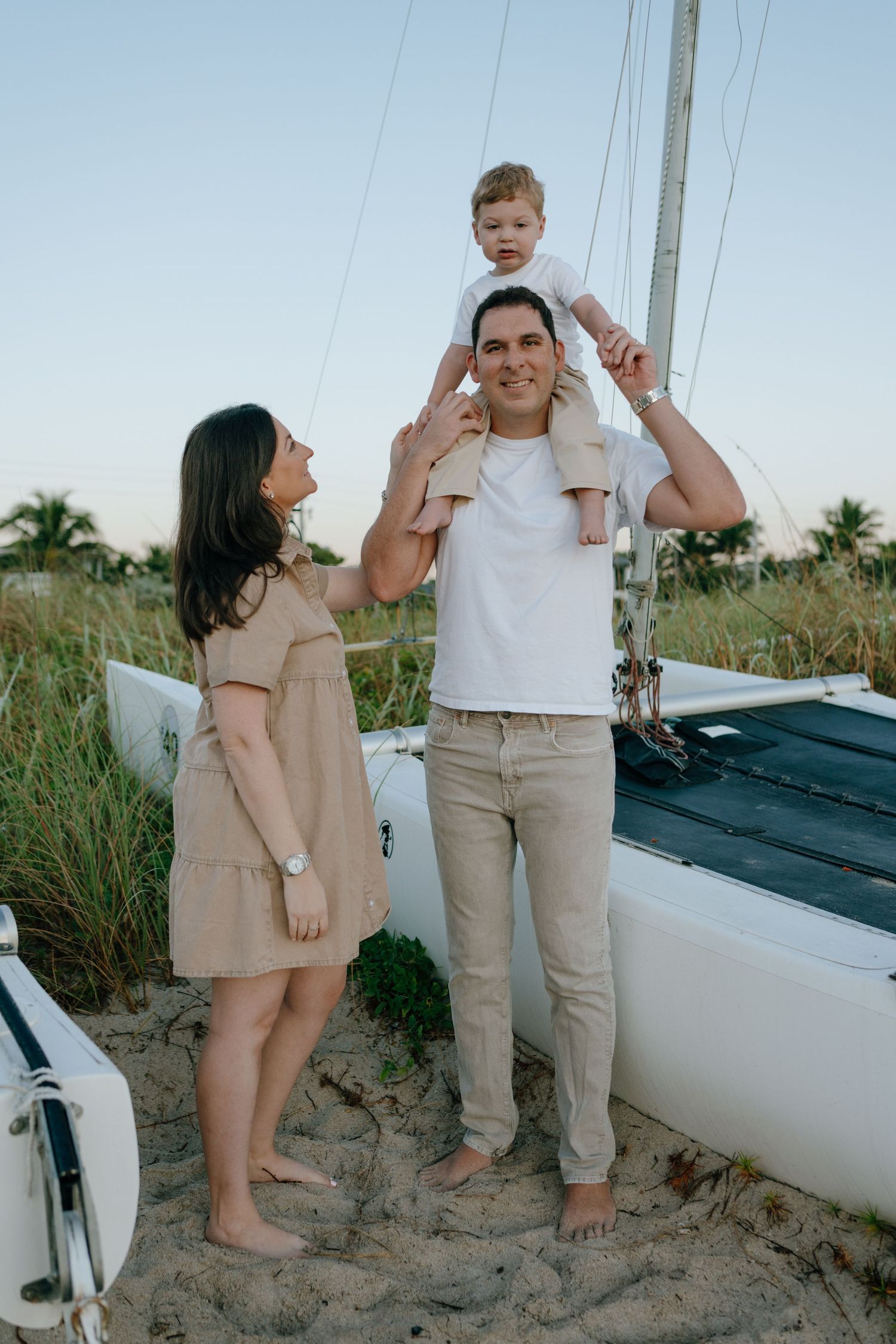 Young family of three posing beside a sailboat on a sandy beach at dusk with palm trees behind.