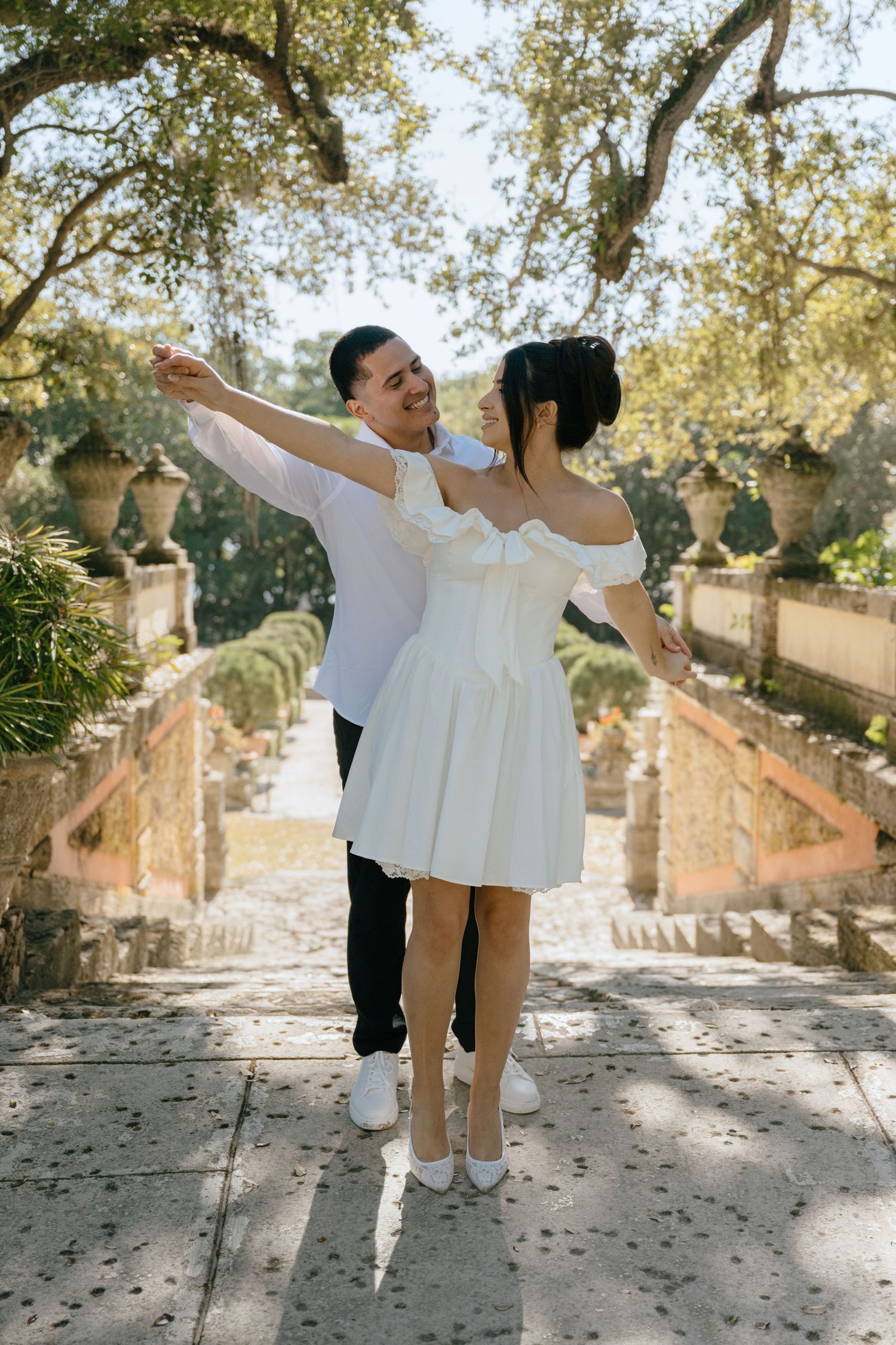 Joyful couple in matching white outfits dance together on a sunlit garden pathway.