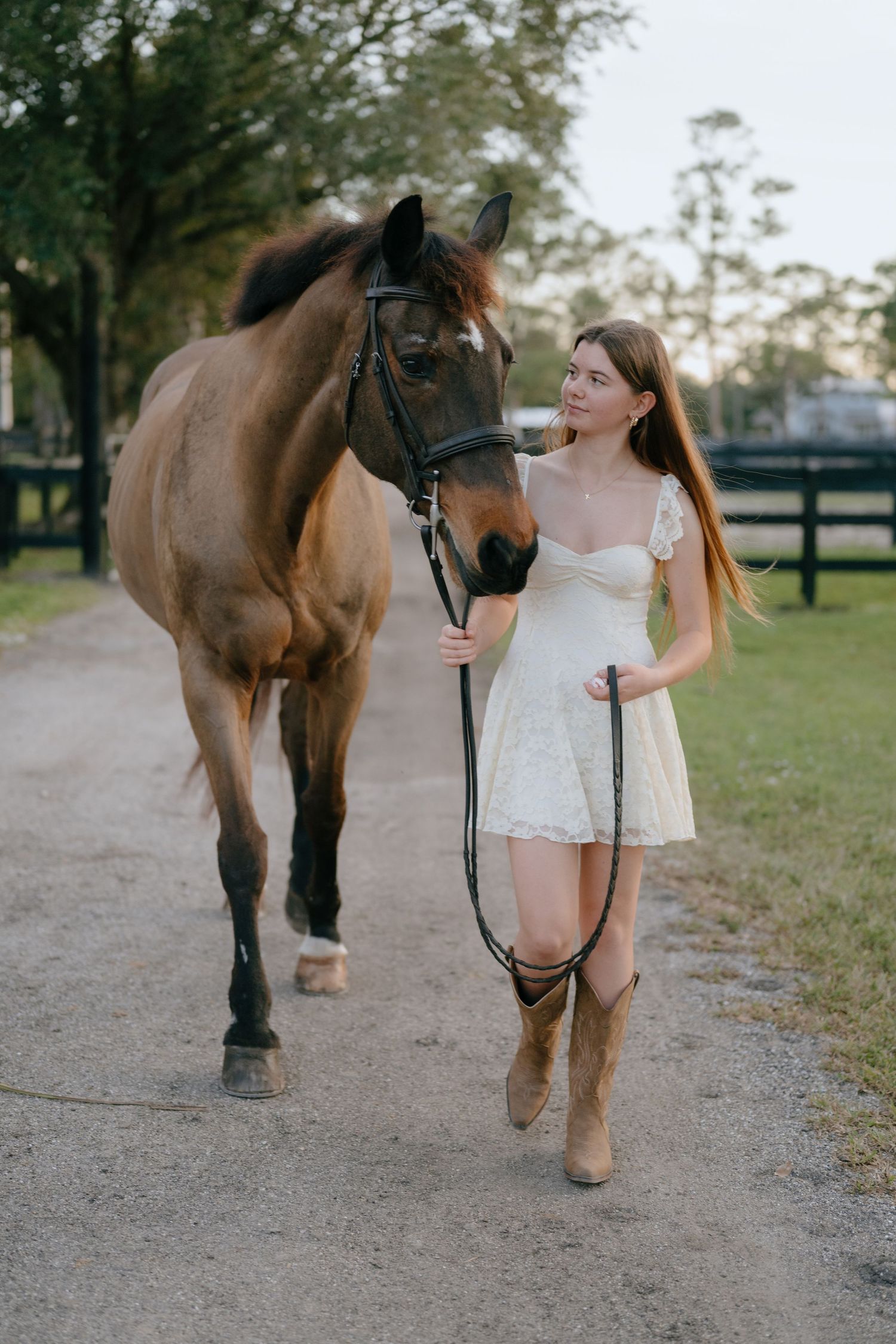 Smiling young woman in white dress and cowboy boots walks a bay horse along a farm path.