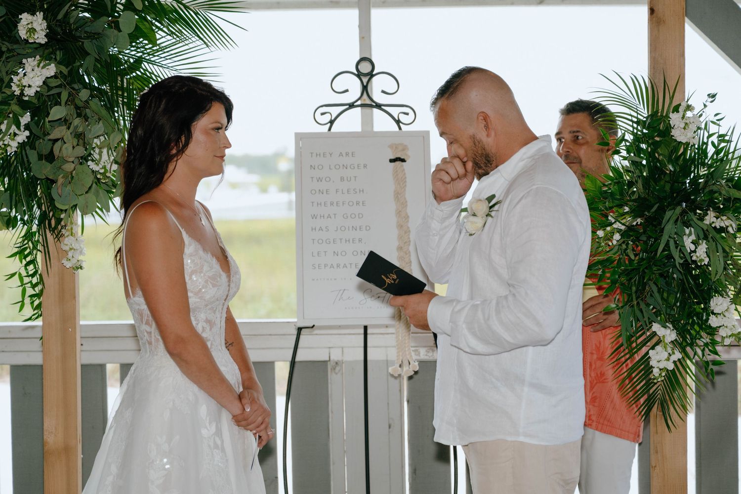 Emotional groom wiping tears during outdoor beach wedding ceremony while bride stands opposite him.
