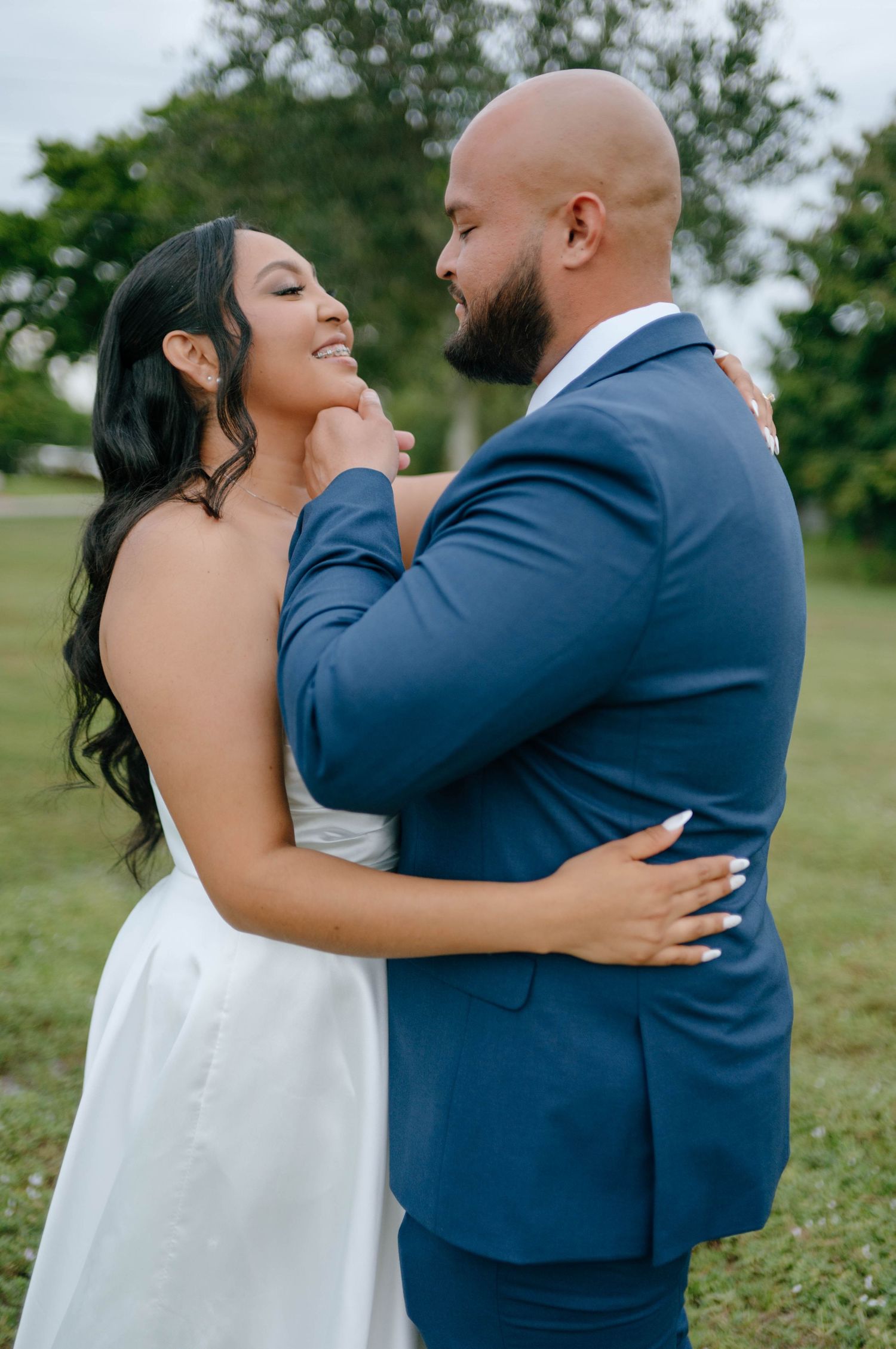 Groom in navy suit tenderly holds bride's face as she smiles joyfully during outdoor wedding portraits.