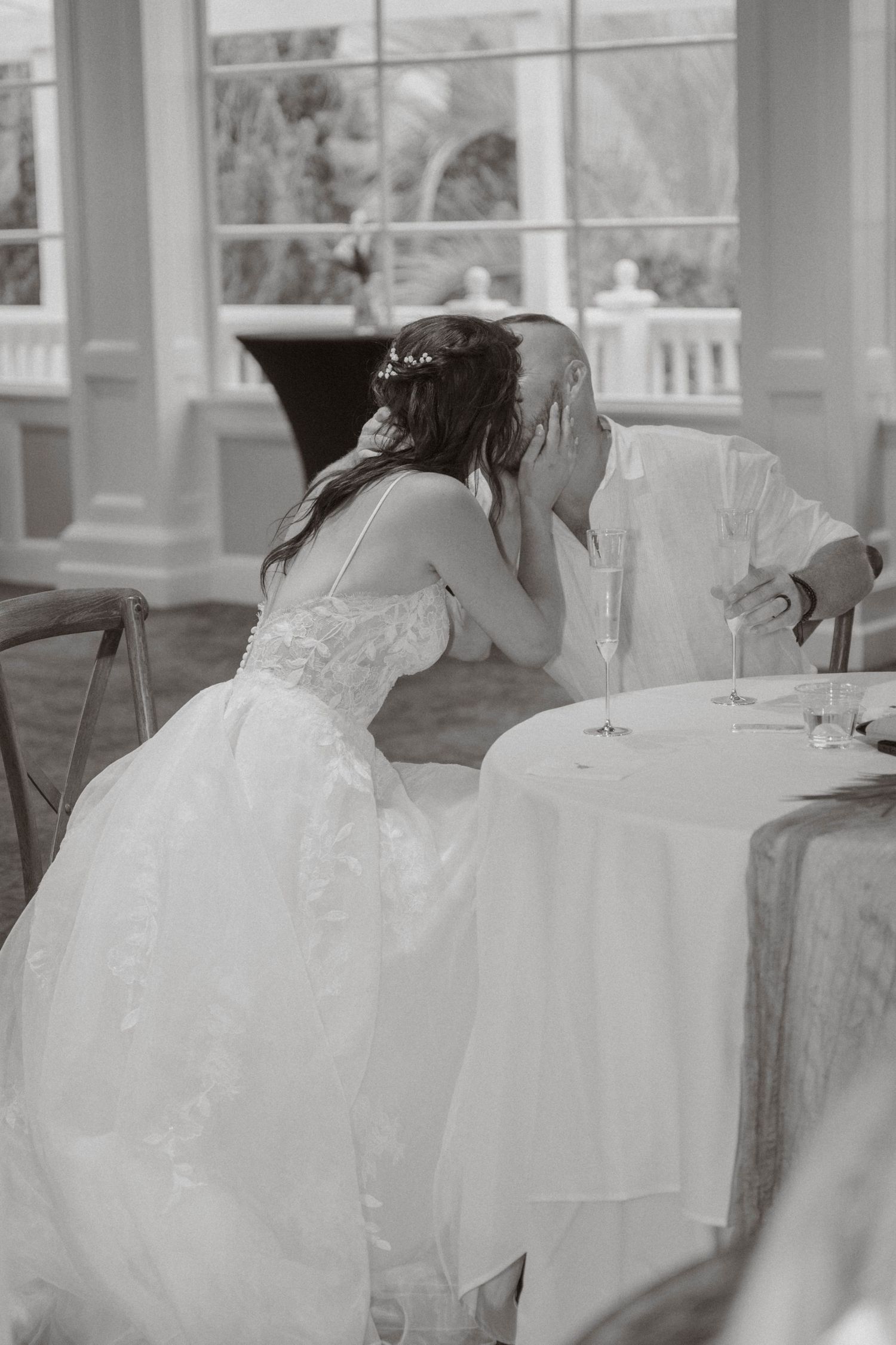 Black and white photo of bride leaning in to comfort emotional groom at wedding reception table.