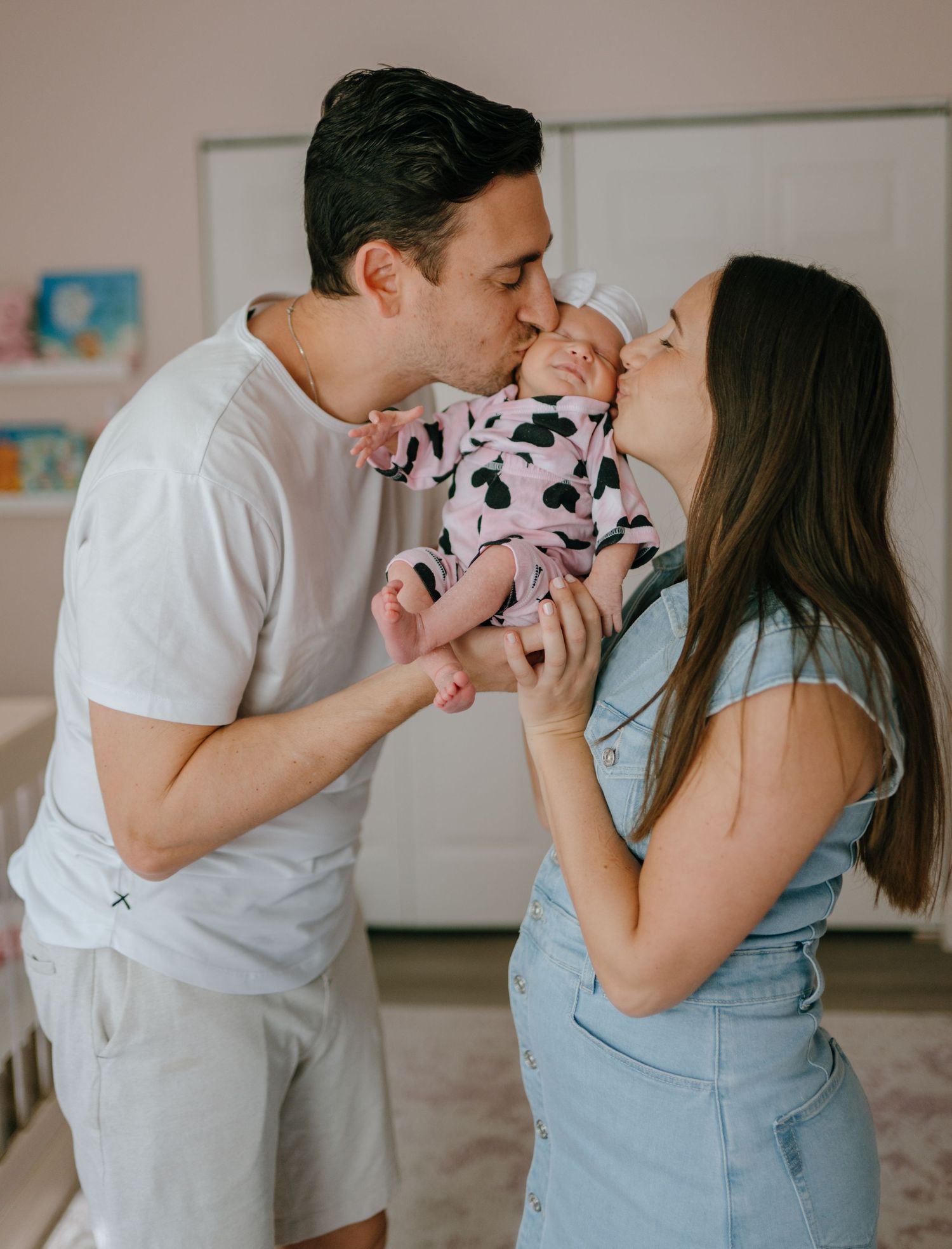 Parents kissing their newborn baby dressed in pink in a softly lit nursery room.