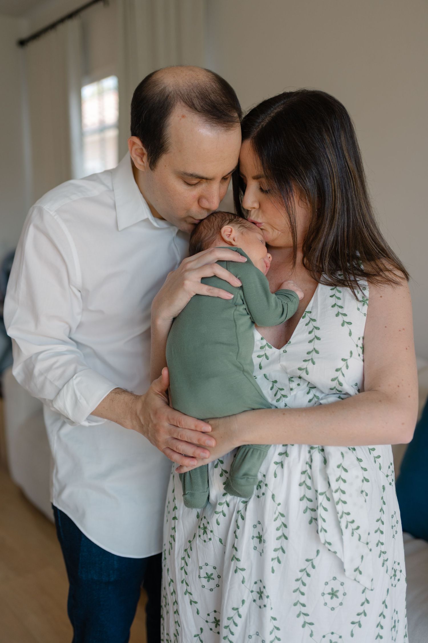 Parents kissing their newborn baby dressed in green in a cozy home setting.