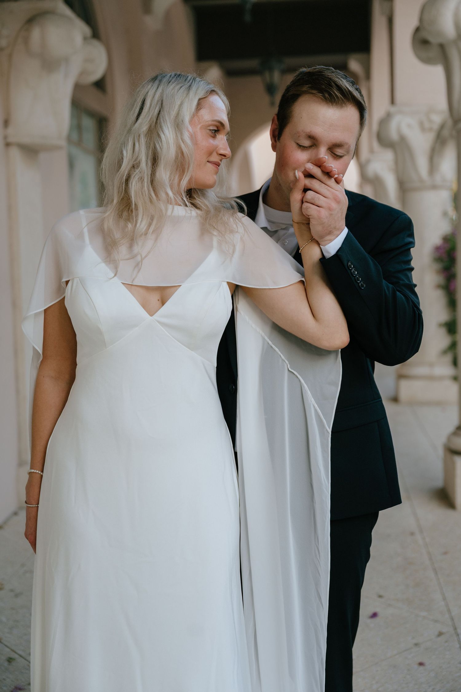 Groom tenderly kisses bride's hand while she smiles, both dressed in elegant wedding attire.
