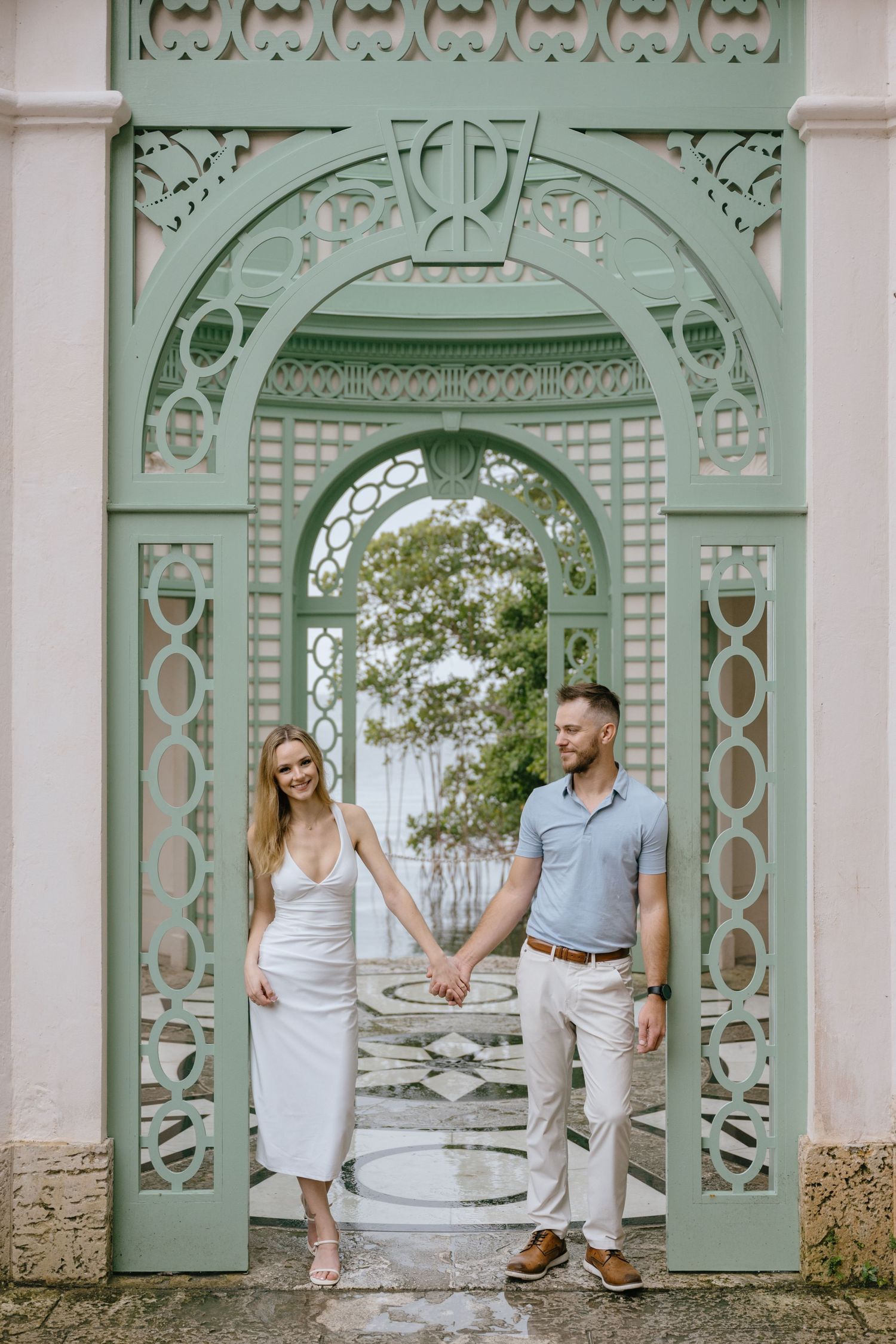 Couple holding hands standing beneath an ornate mint green decorative archway during an engagement photo session.