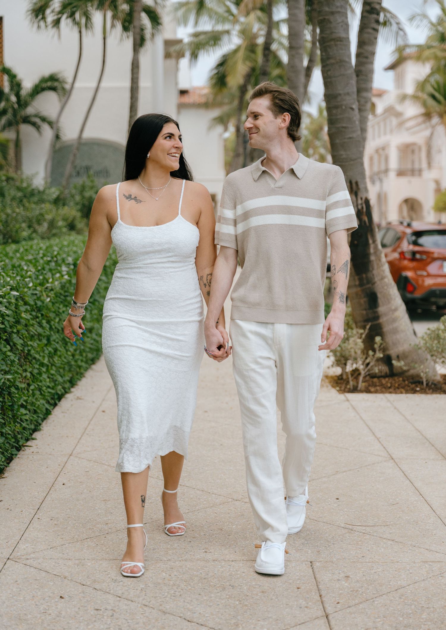 Smiling engaged couple holding hands and walking together along a sunny Palm Beach sidewalk lined with palms.
