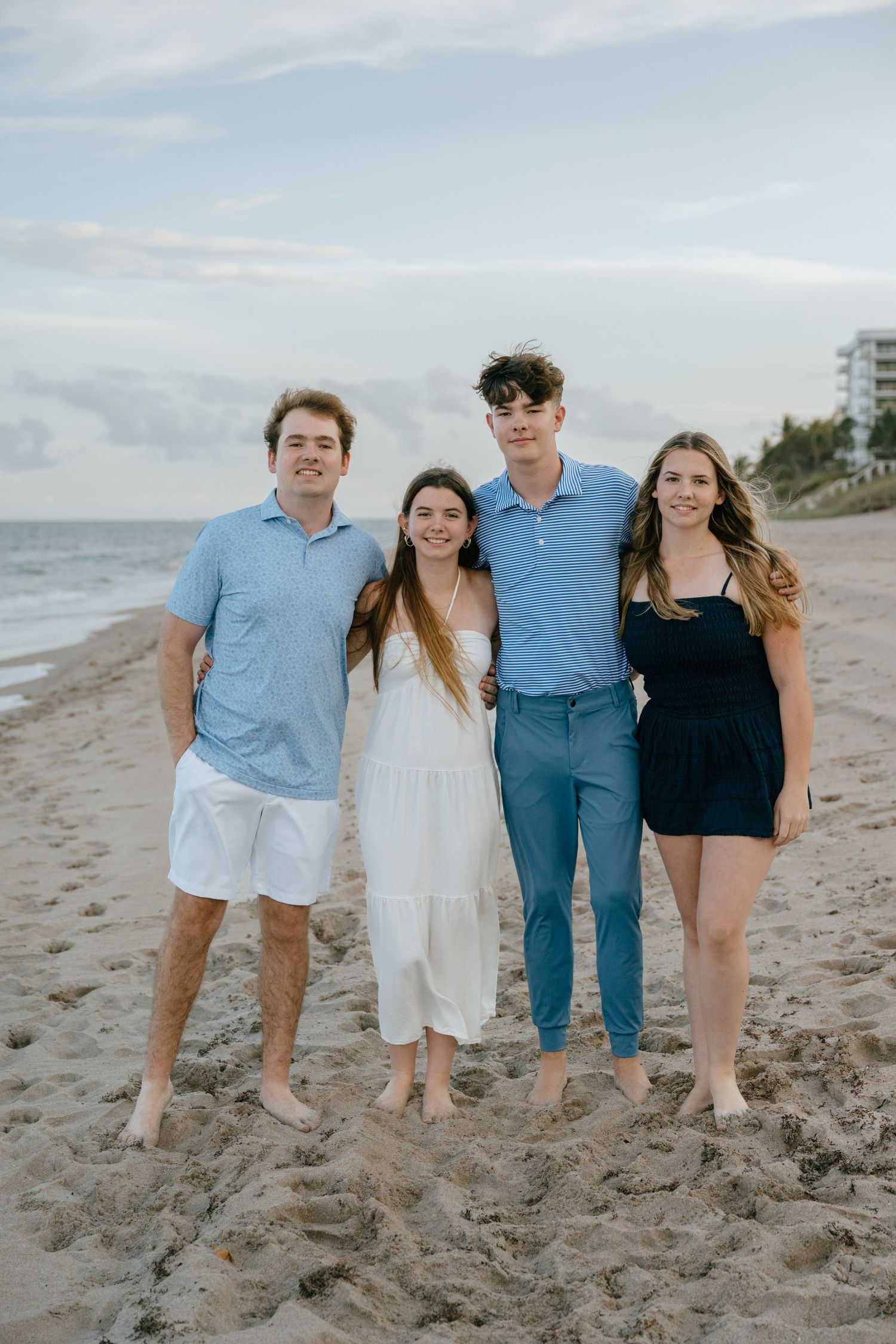 Four young adult siblings posing together on a sunny beach in coordinated blue and white outfits.
