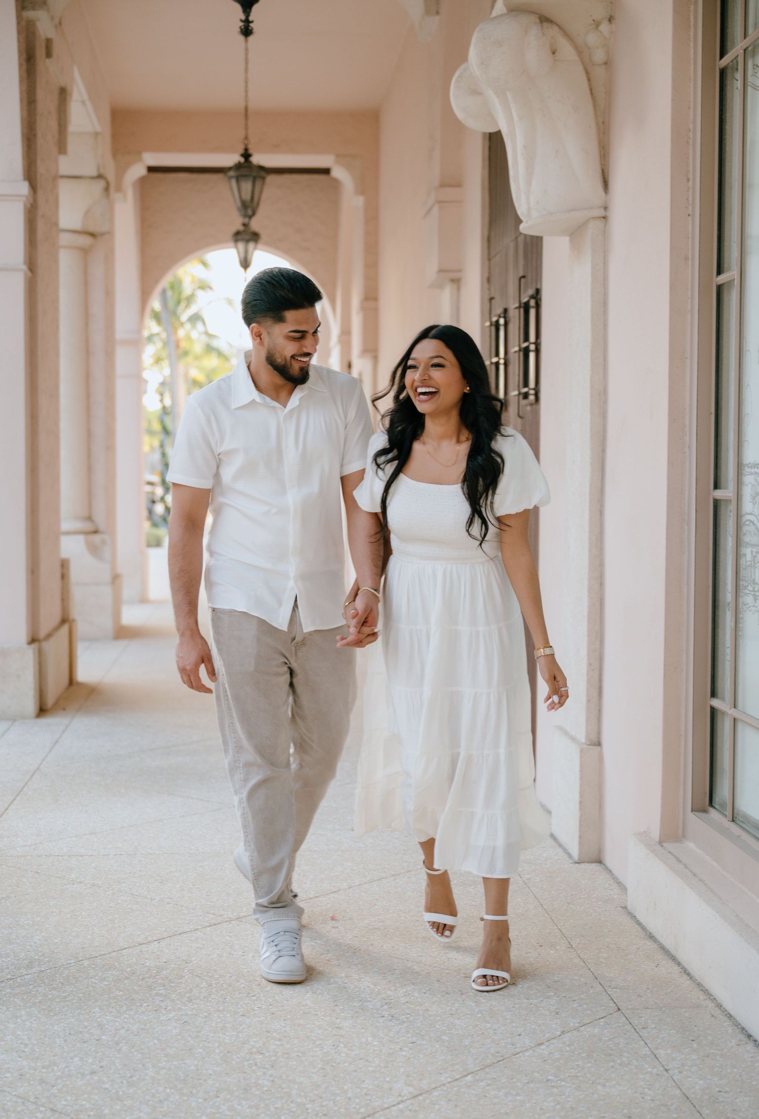 Happy couple in white outfits holding hands walking through elegant pink archway corridor.