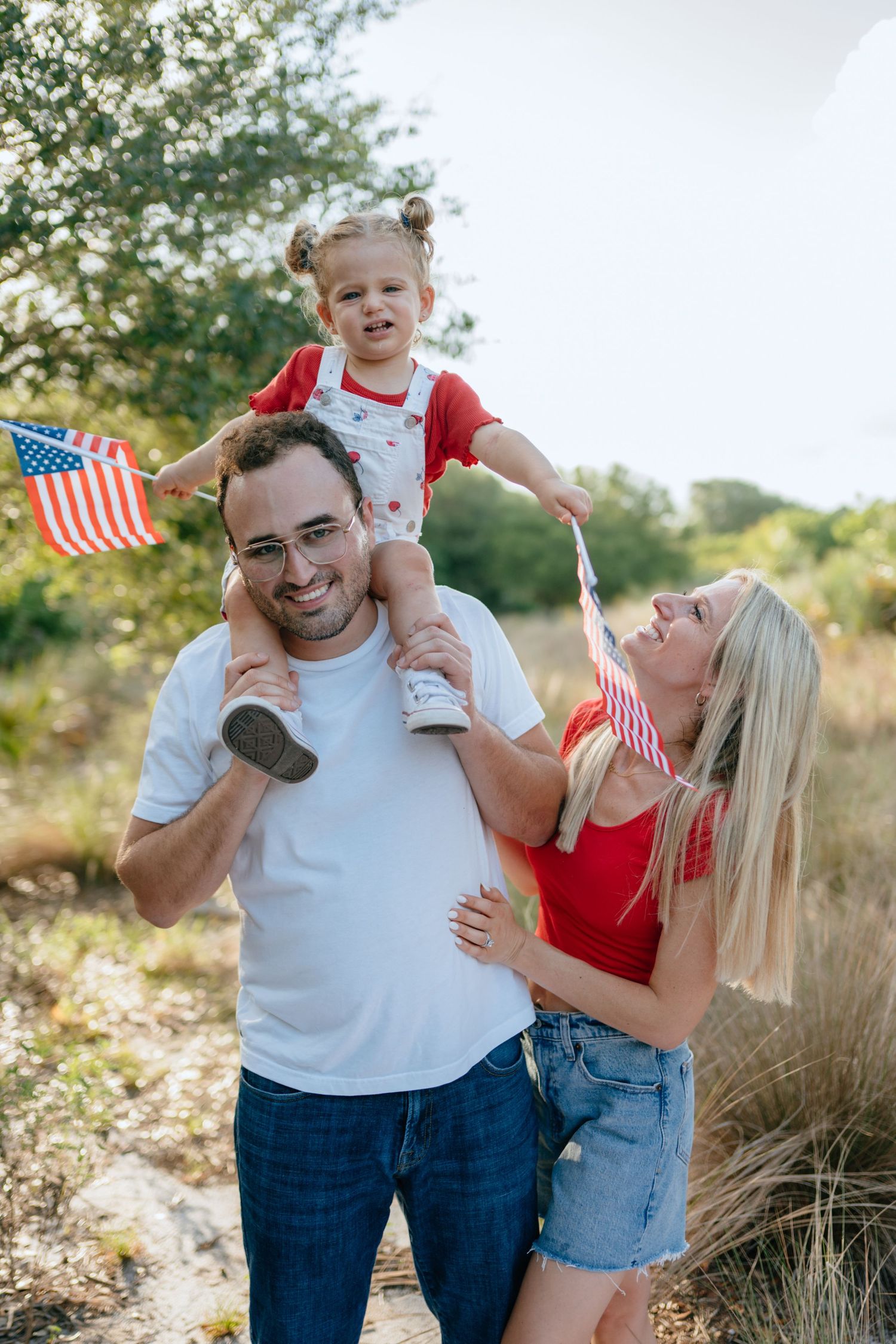 Happy family with toddler on dad's shoulders waving American flags outdoors on a sunny day.