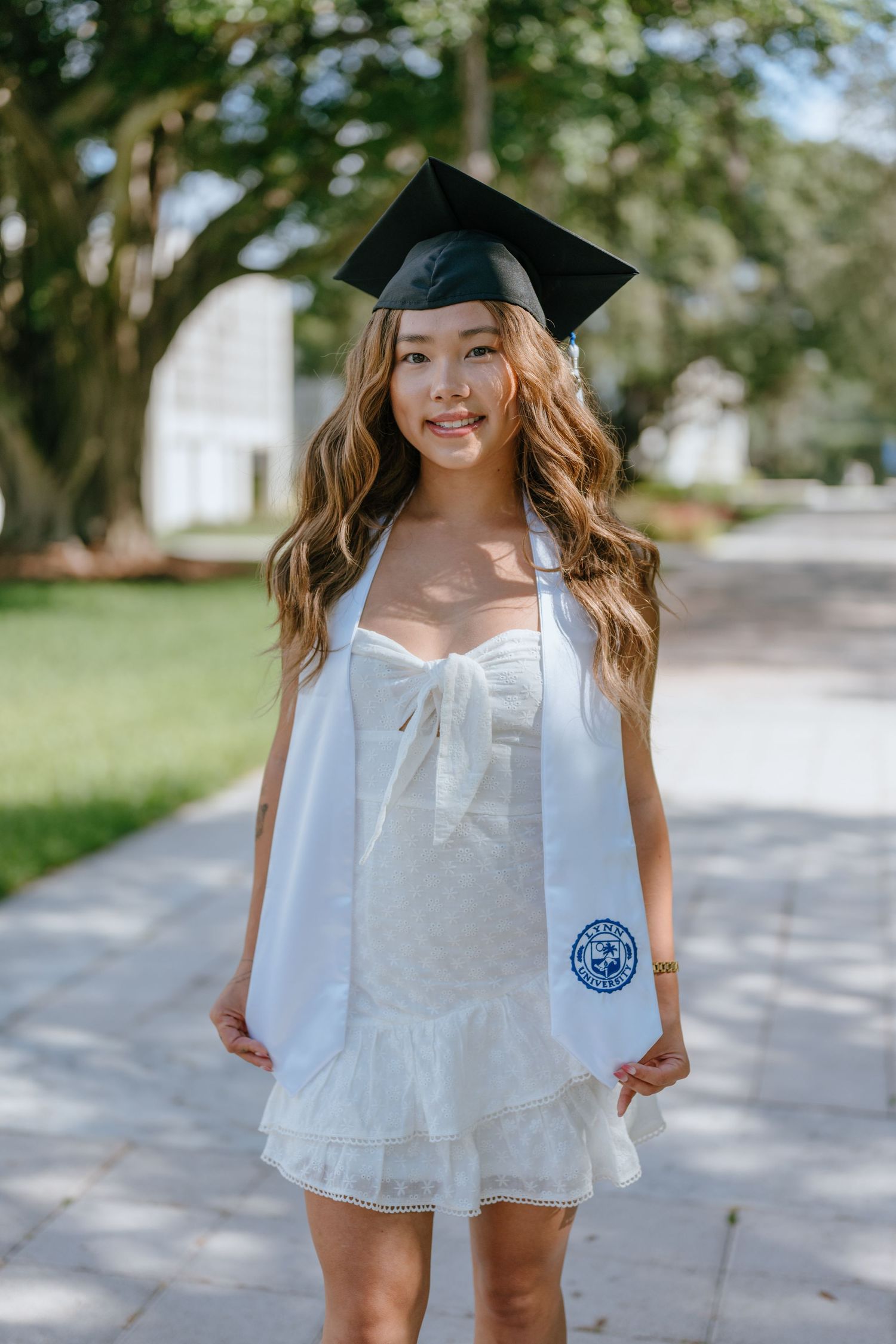 Young woman in white dress and graduation cap with Lynn University stole smiling outdoors.