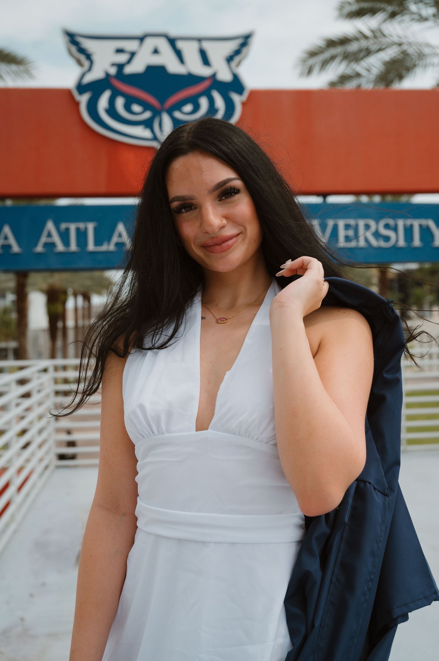 Smiling woman in white dress holds graduation gown over shoulder in front of FAU owl logo sign.