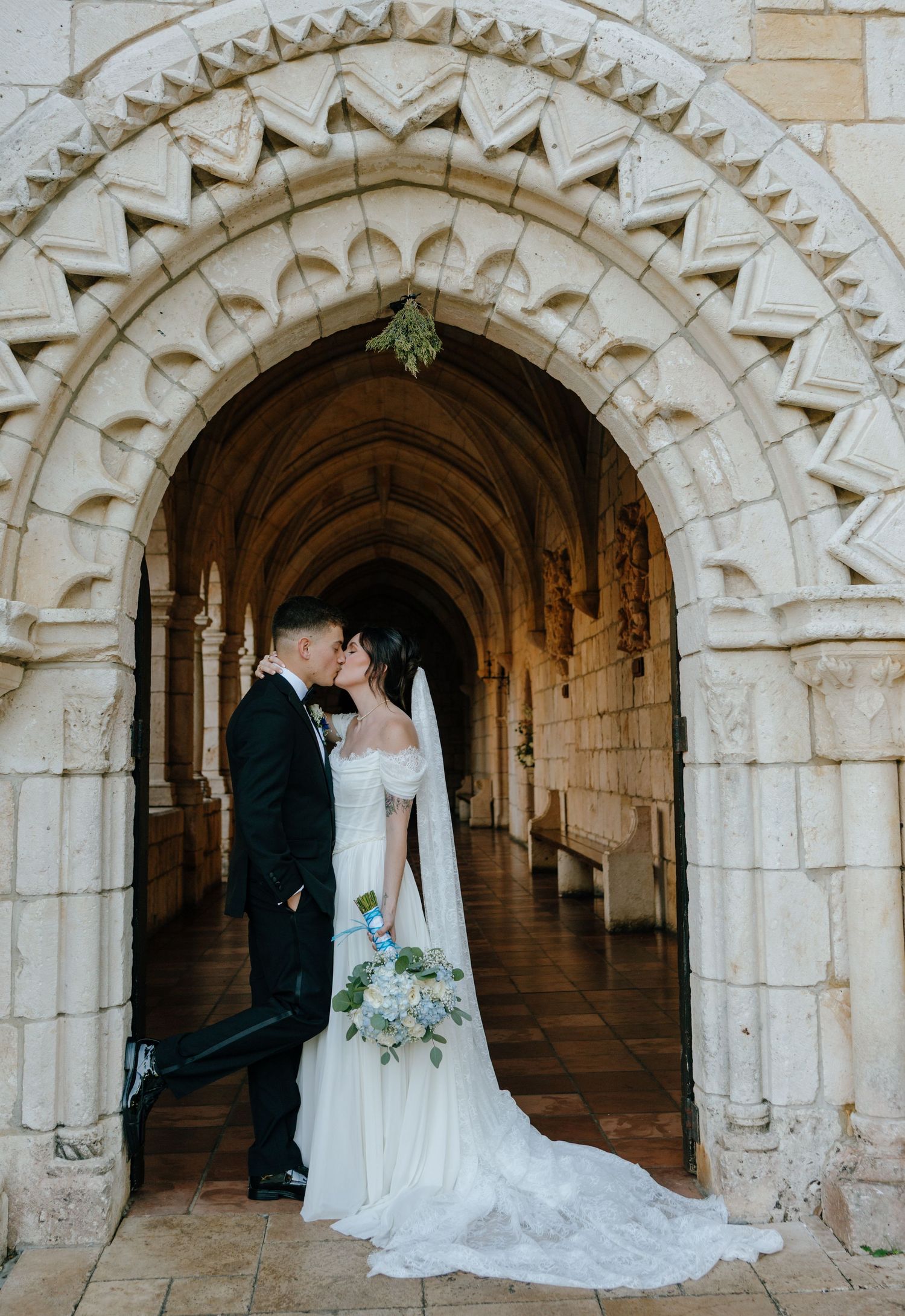 Bride and groom kissing under an ornate stone archway in a historic chapel corridor.