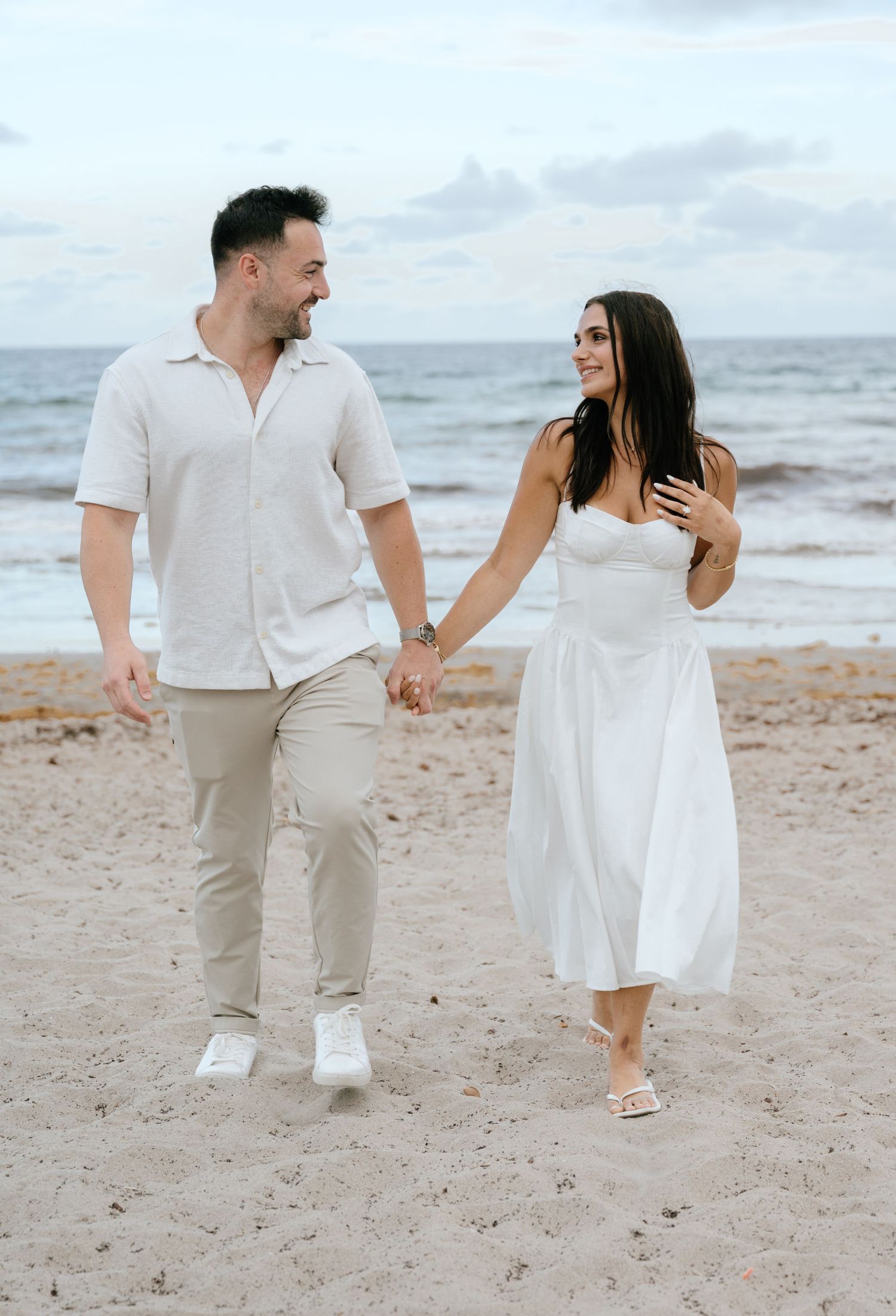 Happy engaged couple walking hand in hand along sandy beach smiling at each other near the ocean.