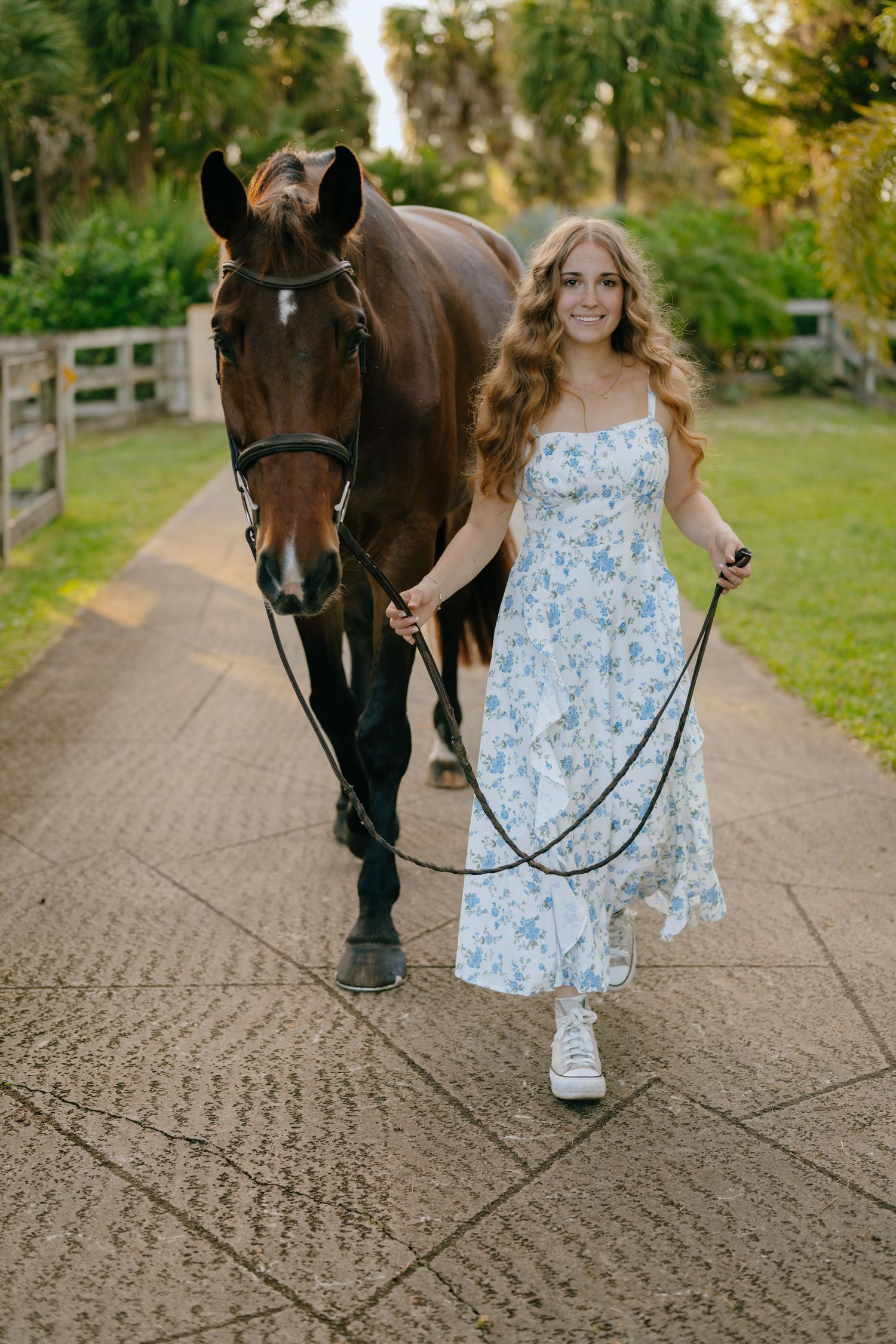 A smiling teenage girl in a blue floral dress walks alongside a bay horse on a sunny paved path.