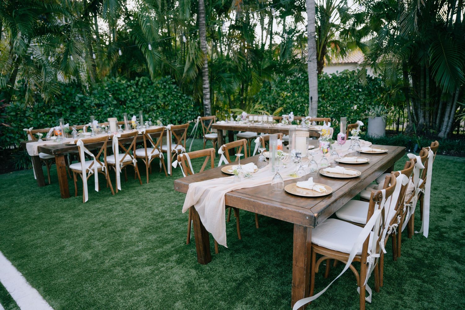 Outdoor wedding reception setup with rustic wooden farm tables and cross-back chairs on green lawn.