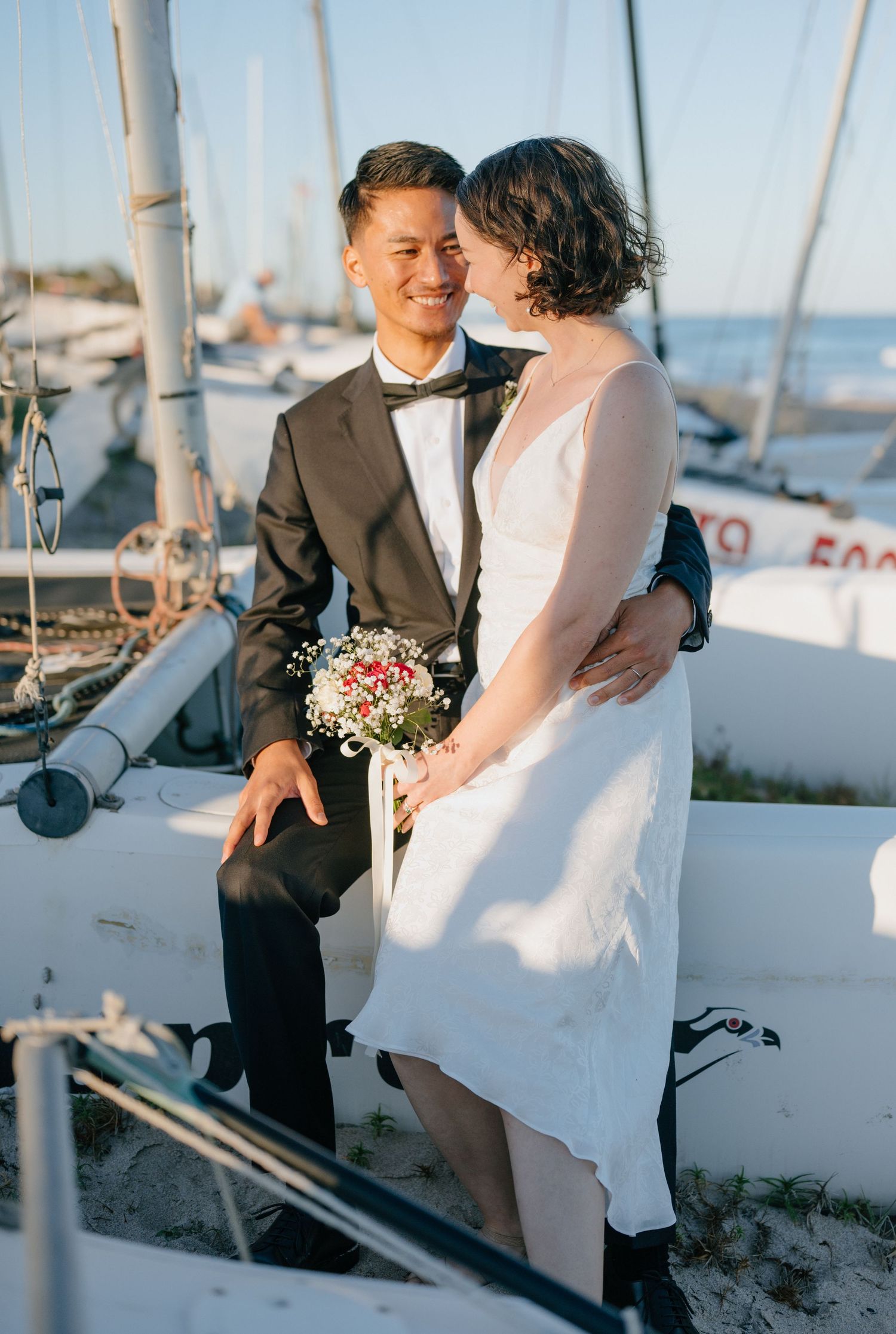 Newlywed couple smiling intimately aboard a sailboat at golden hour, bride holding a floral bouquet.