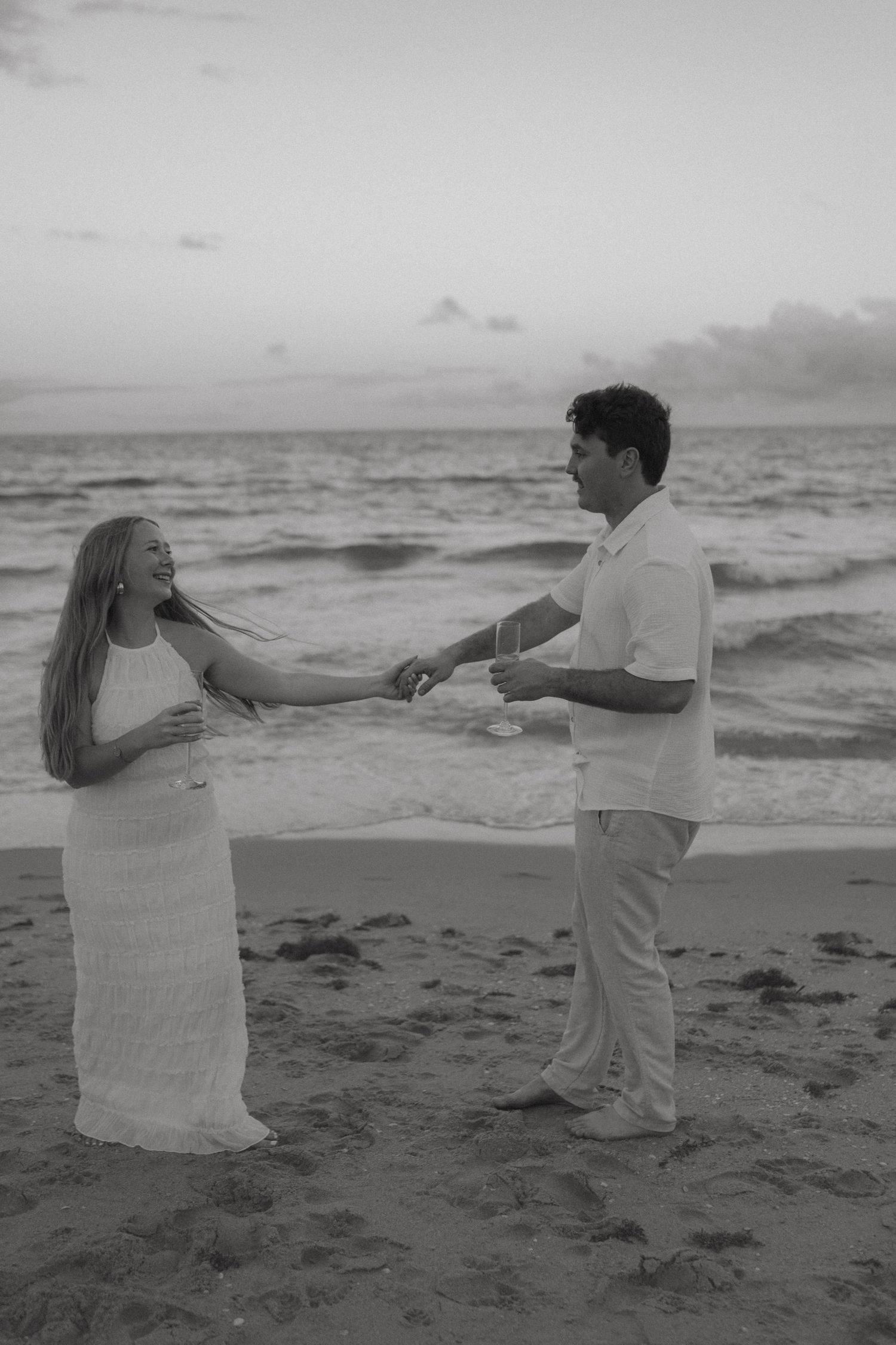 Black and white photo of couple sharing champagne and laughing together on ocean beach.