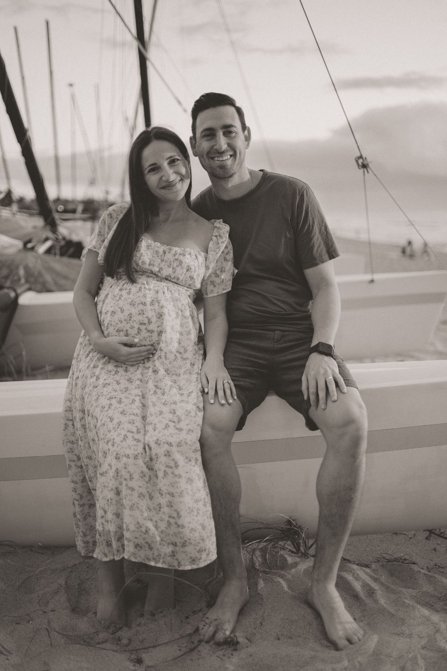 Black and white maternity photo of smiling pregnant couple sitting near sailboats at sunset.