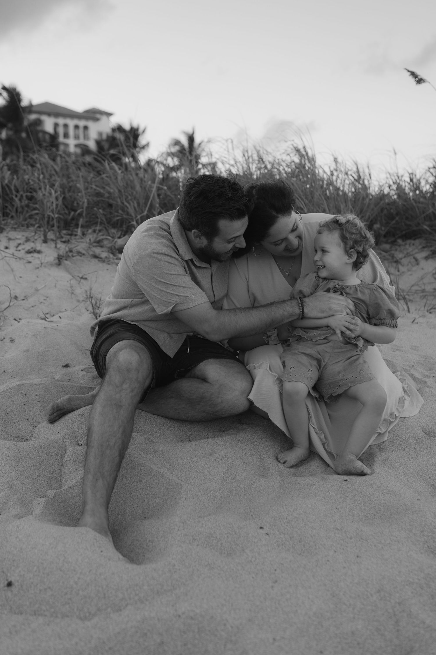 Black and white photo of loving family of three cuddling together on sandy beach dunes.
