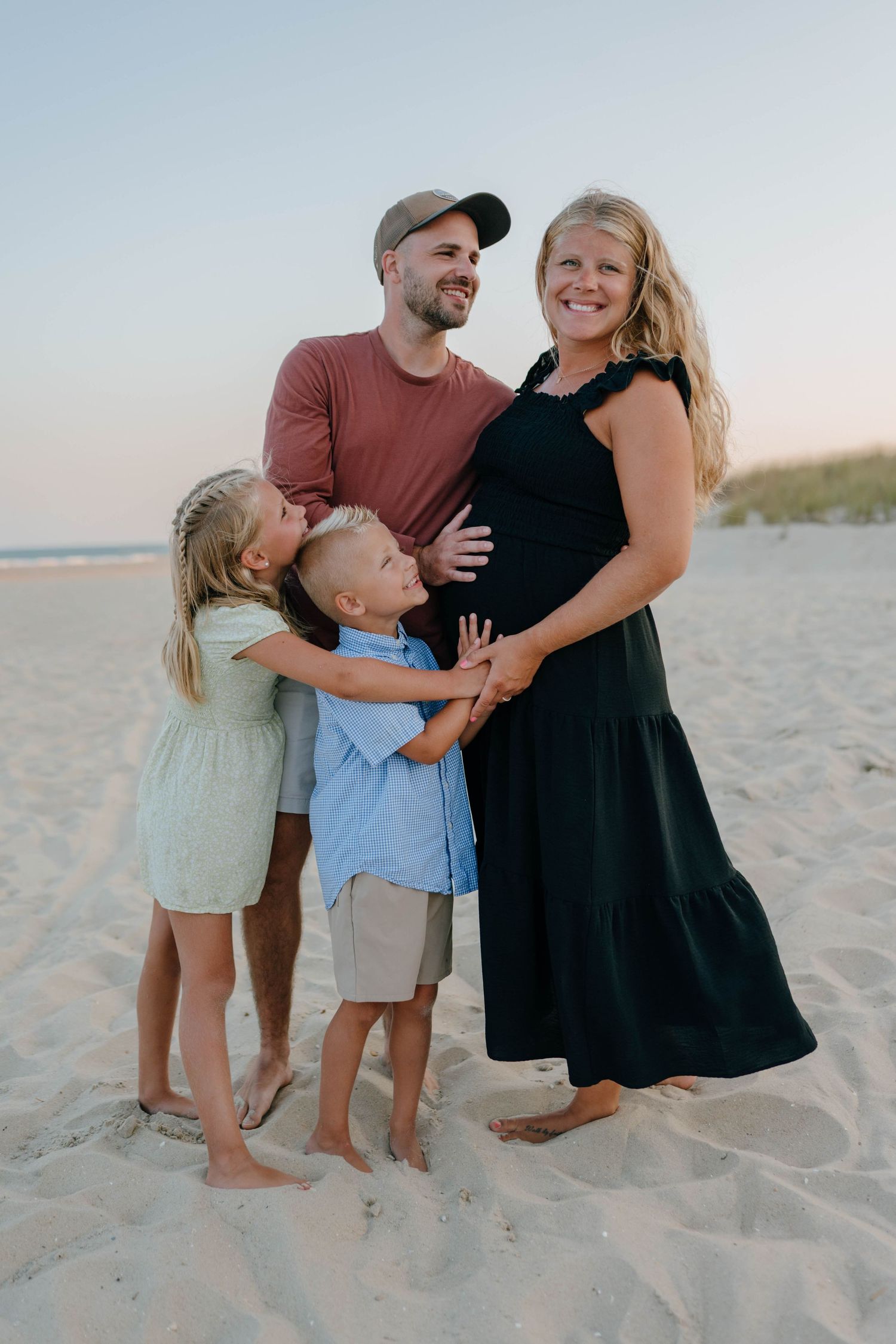 Joyful family beach portrait with children hugging pregnant mothers belly at golden hour on sandy shore.