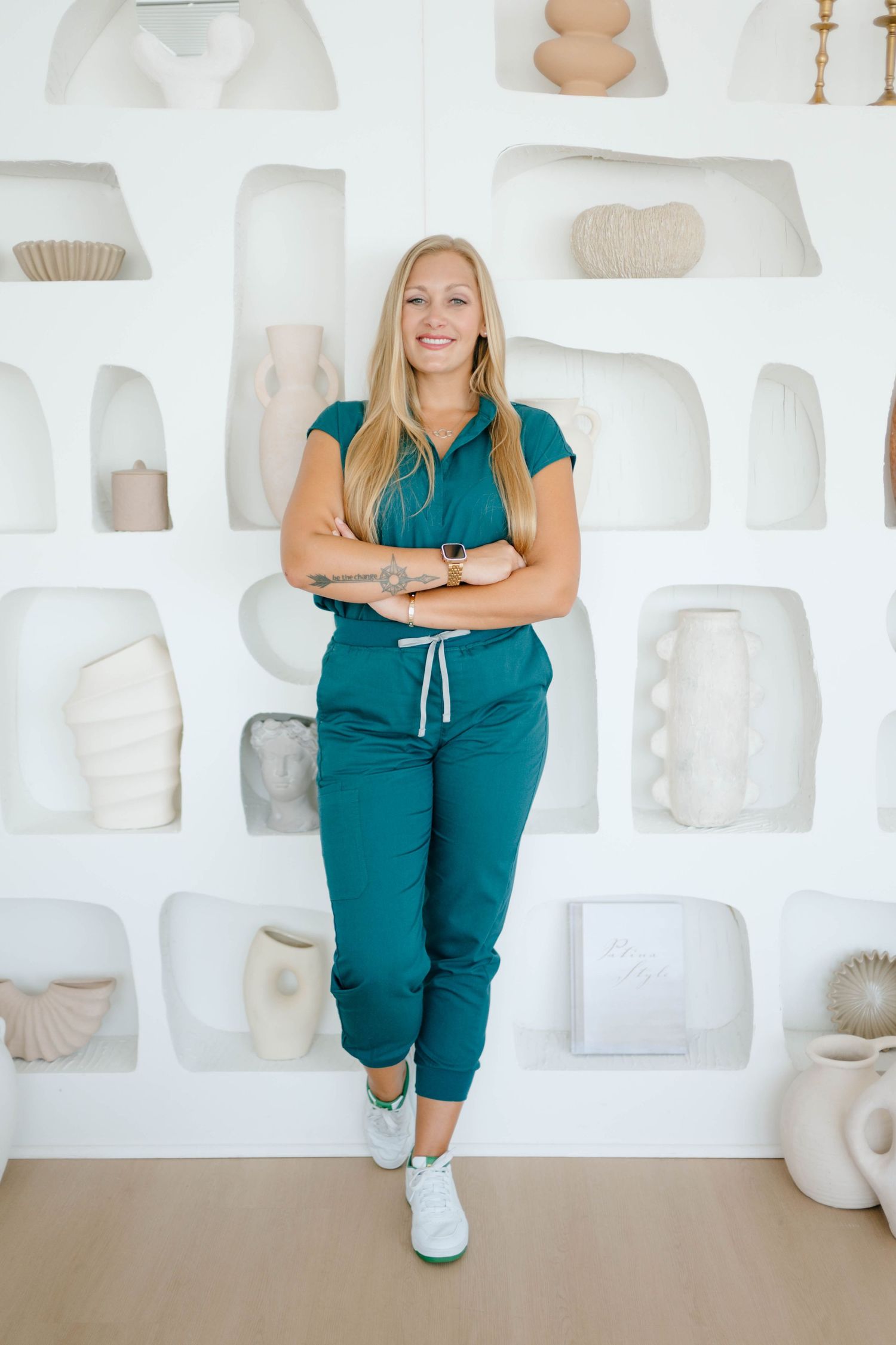 Smiling blonde woman in teal scrubs standing with arms crossed against a white wall with decorative shelving.
