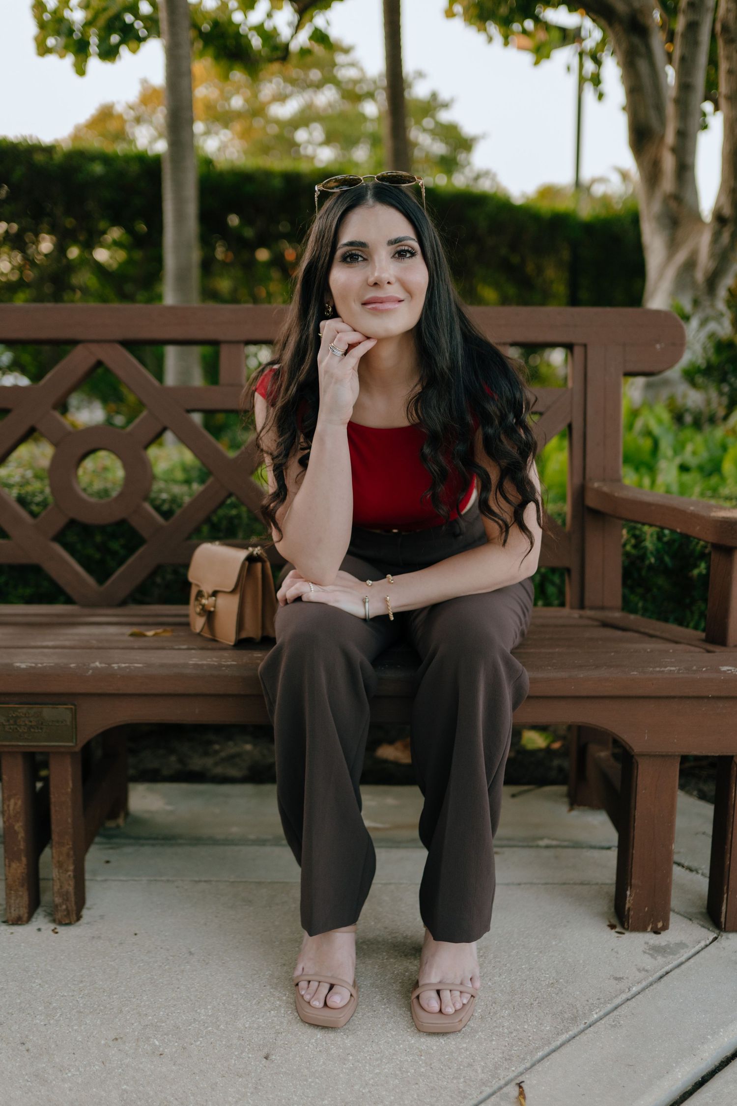 Woman in red top and brown pants sitting on wooden bench in lush garden.